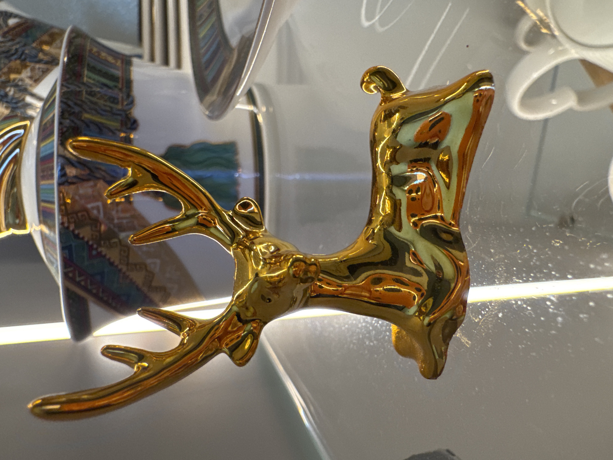 An ornate golden deer figurine rests on a glass shelf, reflecting the ambient lighting. Behind the figurine, a decorative bowl with intricate patterns is visible, along with stacked plates and a white teacup. The overall setting appears to be a retail display or a home environment. The specific location in Sochi, Russia cannot be determined from the provided image.