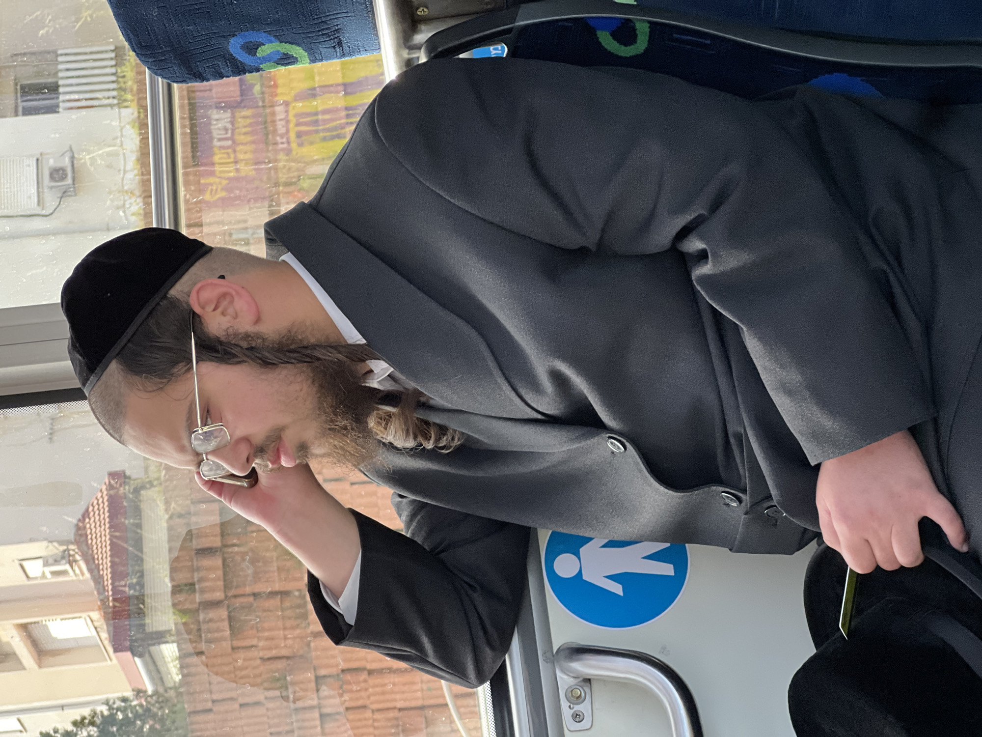 A man with a beard and glasses, wearing a black hat and suit, appears to be sleeping on a bus. He is leaning against the side of the bus, which has a blue and white sign with a pedestrian symbol. The bus window shows buildings typical of an urban area, and based on visual cues, the image may be taken in Kiryat Bialik, Israel. The bus interior includes a blue seat and a silver handrail.