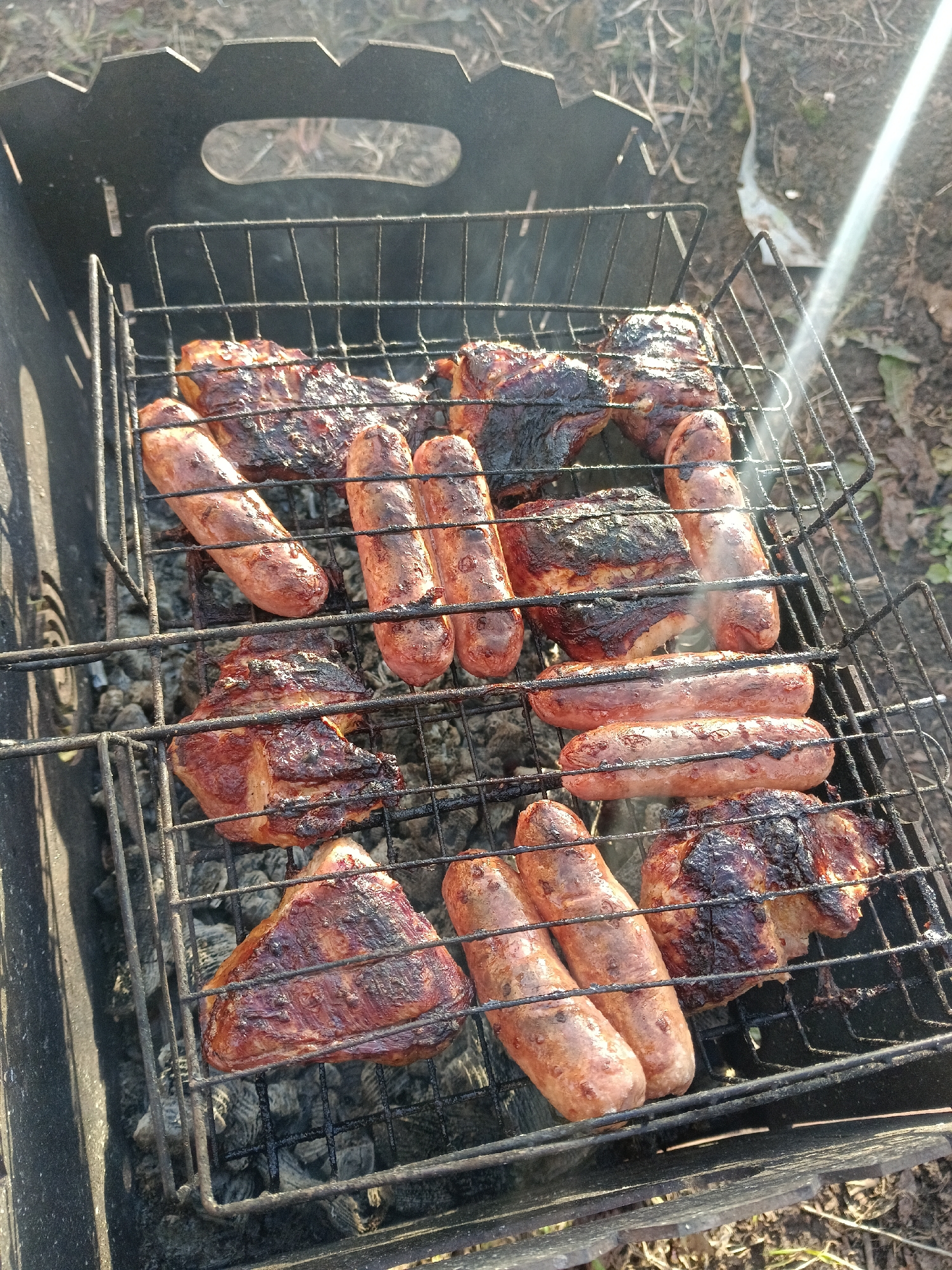 The image depicts a grill filled with sausages and pieces of meat. The food is placed on a metal grate over charcoal within a portable grill. The grilling appears to be taking place outdoors, with some natural ground cover visible around the grill. The image appears to have been taken during the day, based on the lighting. The location is assumed to be Chaikovsky, Russia. There are no people visible in the image.