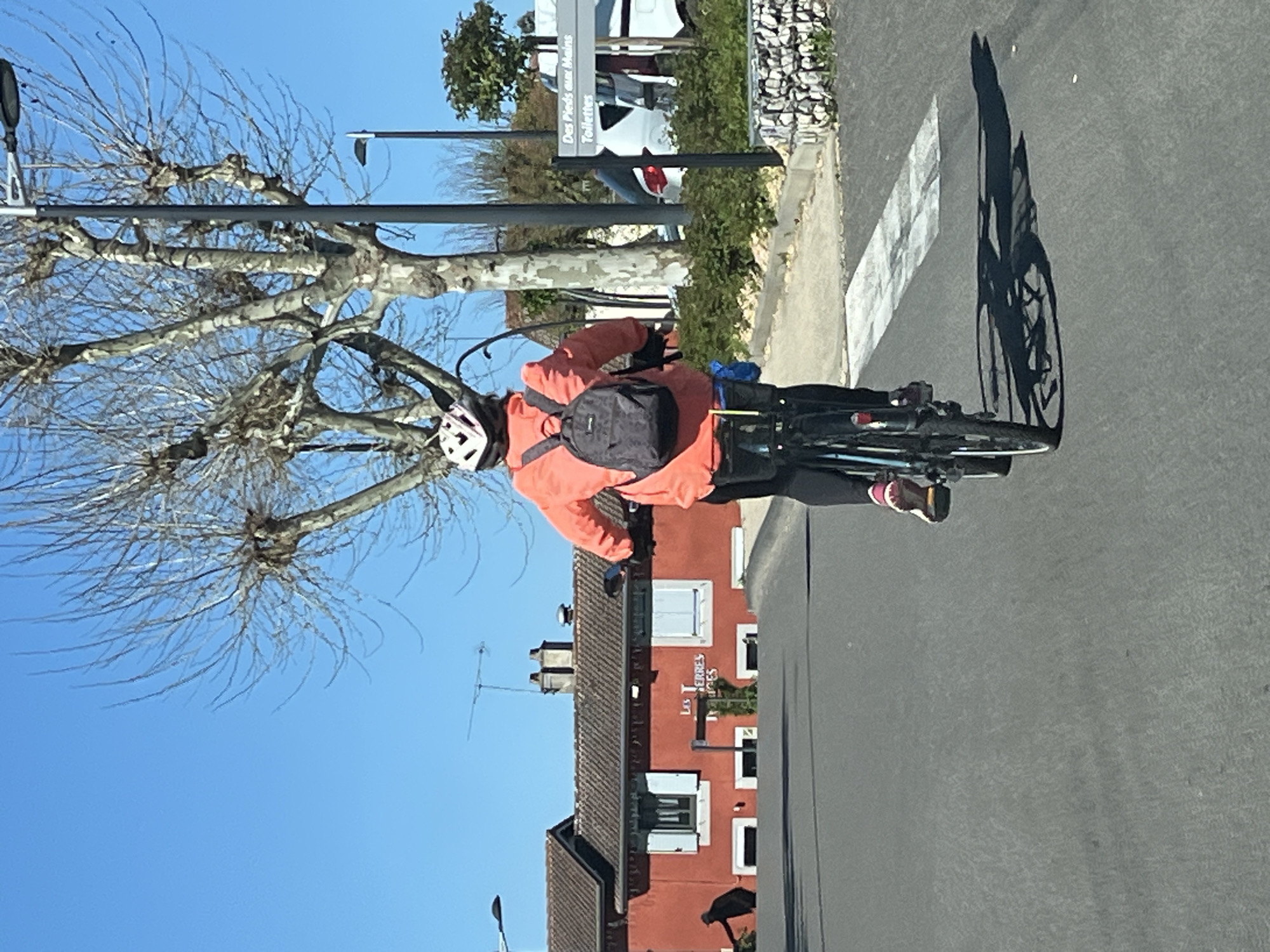 A cyclist rides a bicycle along a road, wearing a helmet, orange jacket, and carrying a backpack. The cyclist is in Chambœuf, France, as indicated by the building's signage "Les Terres Rouges". The weather is sunny, casting a shadow of the cyclist and bicycle on the road. A crosswalk is visible. Bare trees and streetlights are present. Cars are parked near a sign in the background.