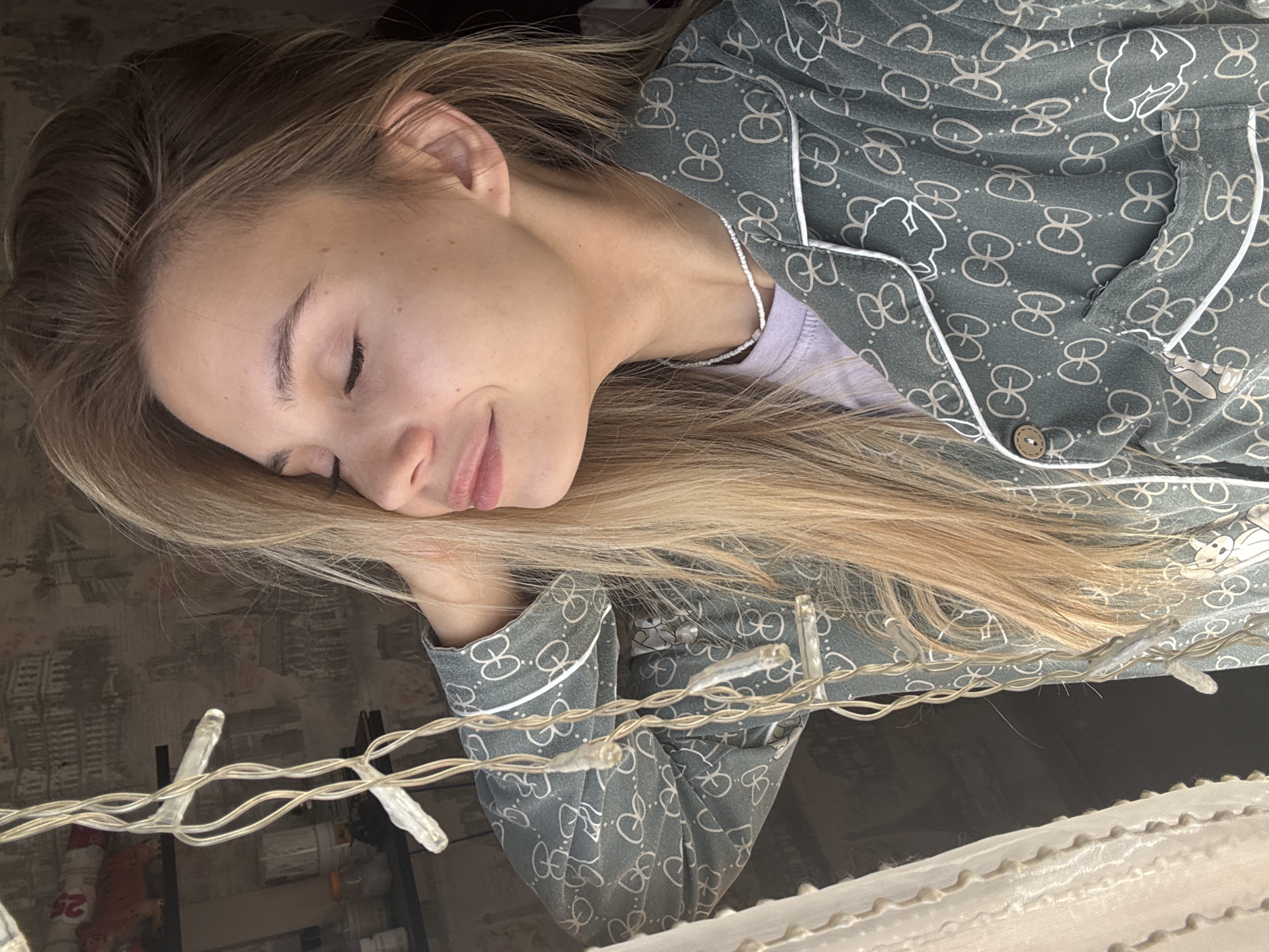 A young woman with blonde hair is pictured from the chest up, against a backdrop of patterned wallpaper. She is wearing a green pajama set with a repeating white logo pattern. Her eyes are closed, and her head is tilted back slightly, with one hand touching her neck. To her left, a string of clear fairy lights hangs in front of a sheer, ruffled curtain. The background wallpaper features an architectural pattern that appears to be buildings or monuments. The overall lighting suggests an indoor setting, possibly a bedroom, during daylight hours.