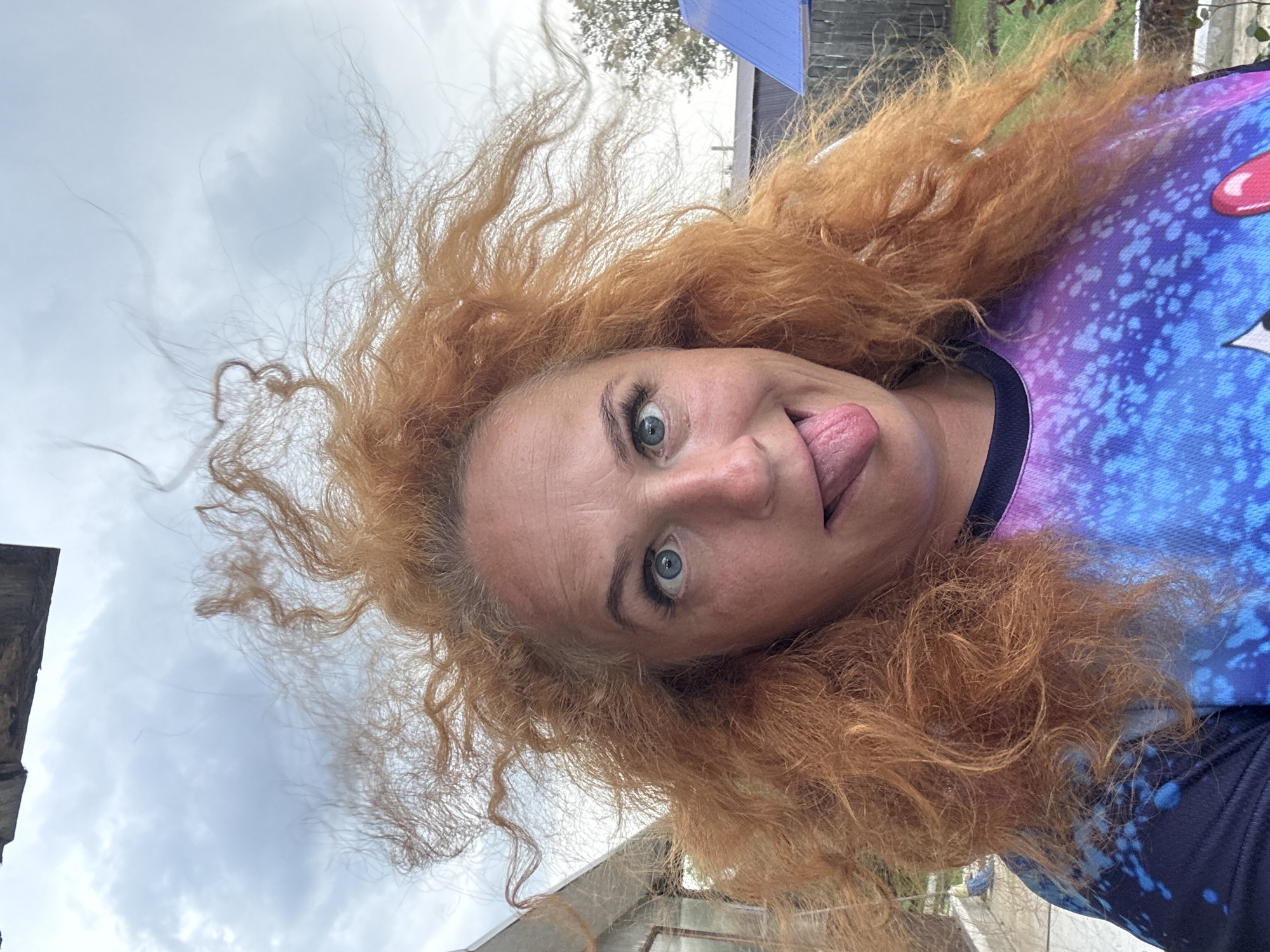 The image shows a close-up selfie of a woman with vibrant, wild red hair against a cloudy sky. She is making a humorous expression, with her eyes wide open and her tongue sticking out. Her face is the central focus, with her hair appearing to be blown by the wind. She is wearing a colorful t-shirt with a galaxy-like pattern of purple and blue. In the background, there are hints of an outdoor environment, possibly a rural or suburban area, with structures that could be part of a house or sheds, and some greenery. The overall impression is one of playful spontaneity. The image is not specific enough to determine its location within the city of Chaikovsky, Russia.