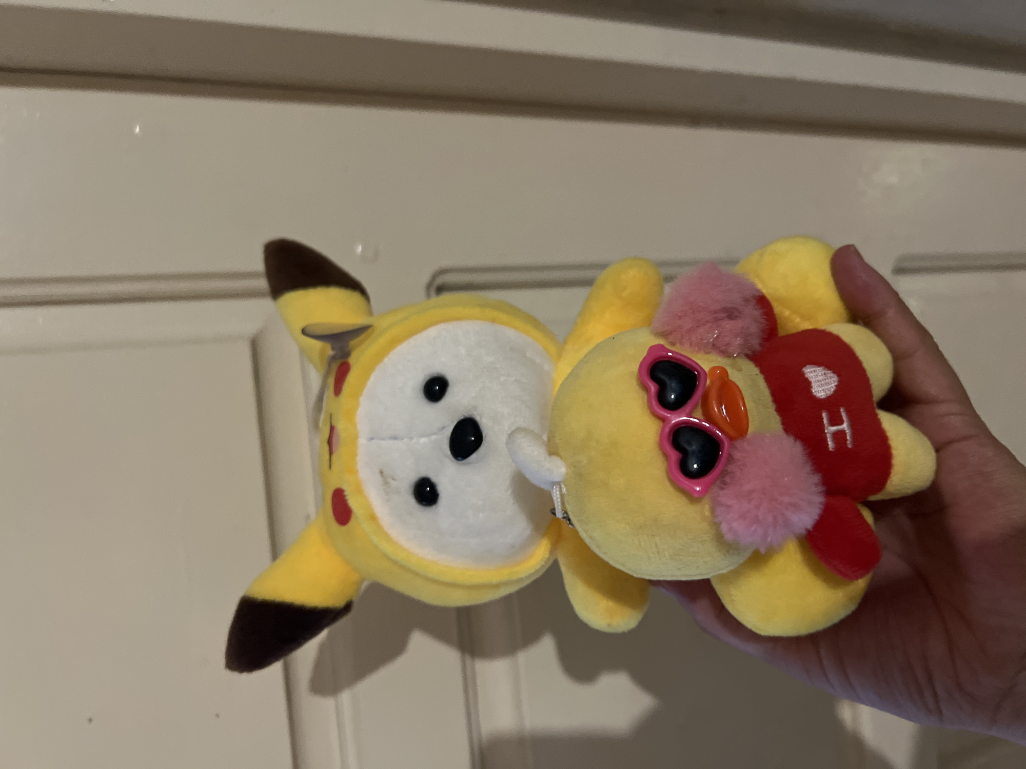 A hand is holding two stuffed toys. The larger toy is yellow with brown accents on its ears and red circles on its head, resembling Pikachu. It has a white face with black eyes and nose. The smaller toy, a yellow duck, is held in front of the Pikachu toy. The duck is wearing pink heart-shaped sunglasses and a red shirt with a white heart and the letter "H" on it. The background appears to be a closed door with vertical panels. The scene is indoors and lit by artificial light, casting shadows on the door. There are no discernible landmarks or indications of the specific location within Bishkek City, Kyrgyzstan.