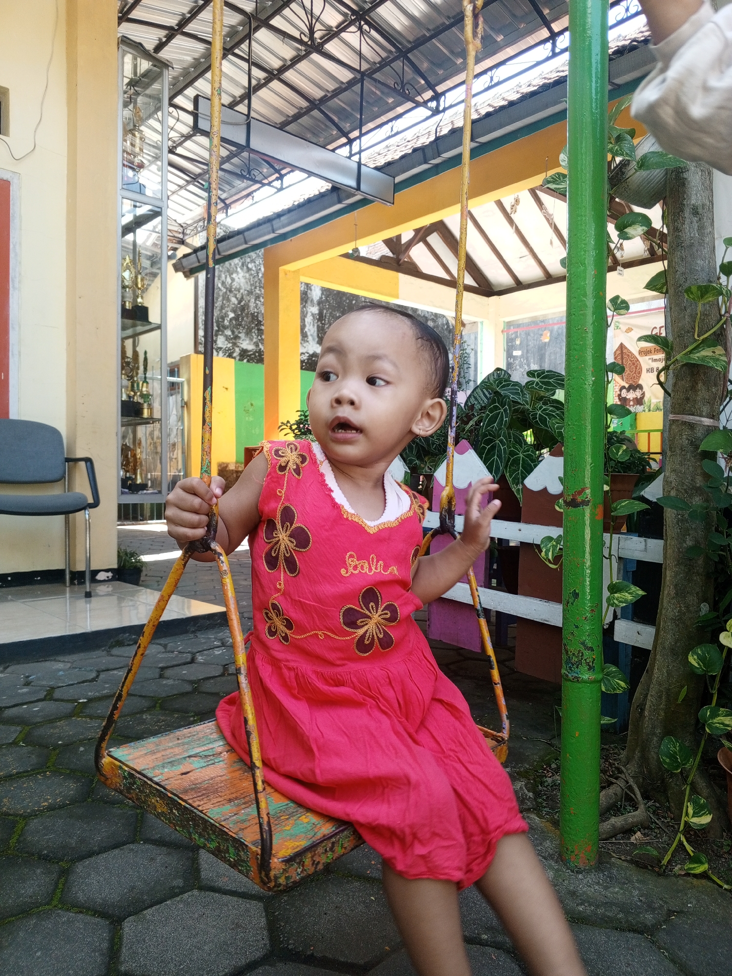 A young child, appearing to be about two years old, sits on a colorful, rustic swing. The child wears a bright pink dress adorned with dark brown floral patterns. The swing set has a weathered wooden seat with traces of orange and green paint, suspended by yellow metal chains from a yellow pole. The child's right hand grips the chain. The background suggests an outdoor setting, possibly a school or public area, with a building visible to the left, painted in yellow and cream. Behind the swing, there's a green metal pole and a tree with lush green leaves. The ground is paved with hexagonal gray stones. In the distance, a rack of trophies can be seen through a glass-paneled section of the building. The image appears to be taken in Sumbersari, Indonesia, based on the context provided.