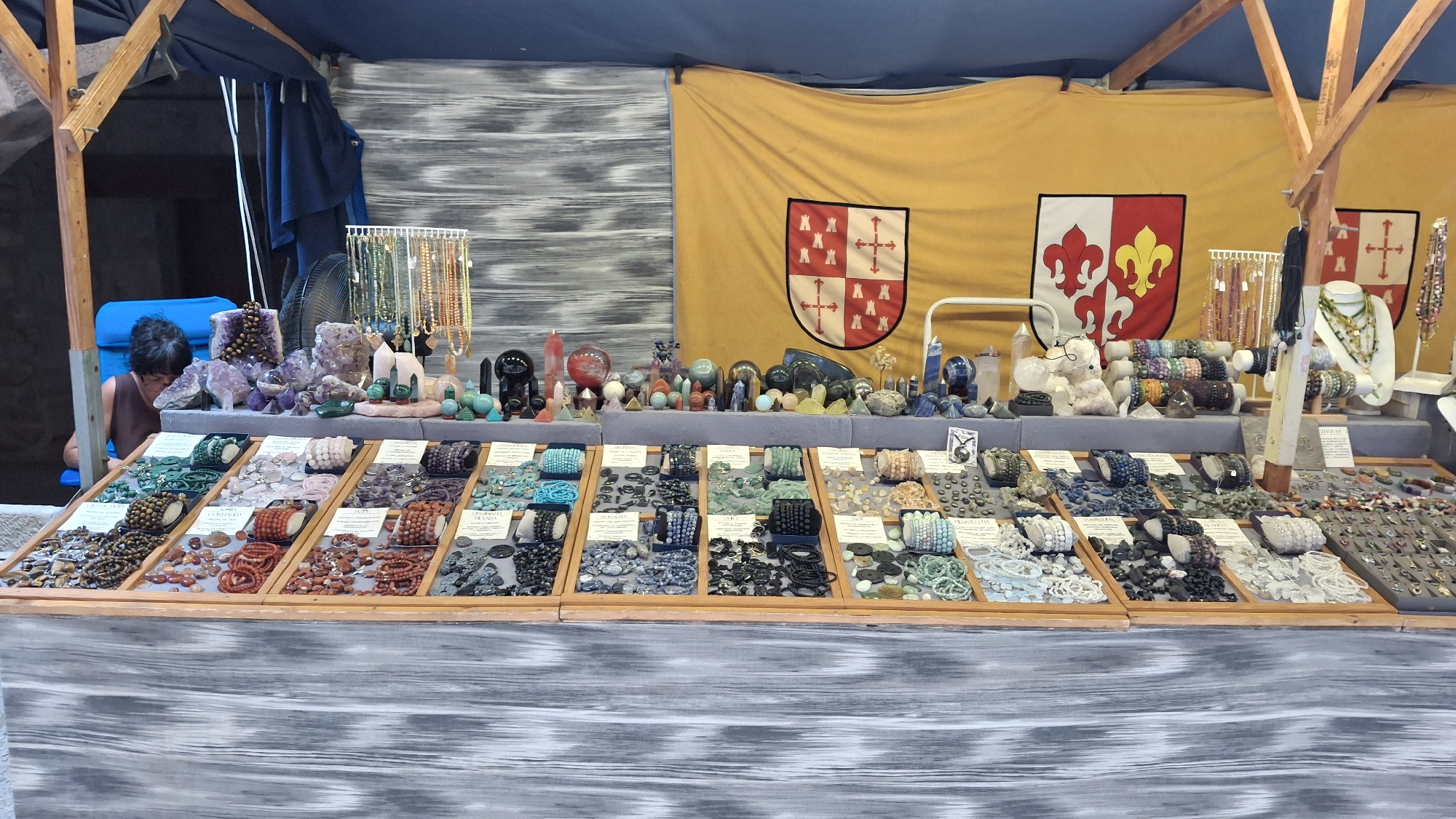 The image depicts a stall at an outdoor market, likely a craft or gemstone fair, in La Fresneda, Spain. The stall is laden with a diverse array of handcrafted jewelry, primarily bracelets and necklaces, and decorative crystal formations. The items are arranged in wooden display trays with individual compartments, showcasing a variety of stones and beads in different colors and textures.

In the background, a large yellow banner serves as a backdrop, featuring two heraldic shields. The stall itself is framed by wooden supports and a dark blue awning overhead, suggesting a temporary structure common at outdoor markets. Some of the display items include amethyst clusters, polished spheres, and various beaded necklaces and bracelets, with labels indicating prices or types of stones.

To the left, a person is partially visible, seated behind the stall and looking down at the merchandise, suggesting they are the vendor or an assistant. The overall environment appears to be a sunny day, with the lighting suggesting it is daytime. The stall exhibits a wide range of colors, from the earthy tones of many of the gemstones to the bright yellow of the banner. The display is neatly organized, indicating a careful presentation of goods for sale.