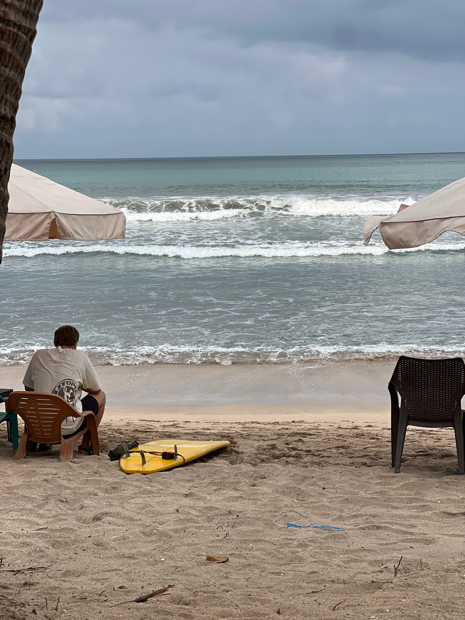 An overcast day at Kuta Beach, Bali, Indonesia. The sandy shore stretches to the horizon, with gentle waves rolling in from the turquoise ocean. Two beach umbrellas, one tan and one lighter beige, are set up on the sand, offering shade. In the foreground, a person wearing a white t-shirt and black shorts sits facing the ocean in a brown plastic chair. Next to the chair, a yellow surfboard lies on the sand. To the right, another dark brown plastic chair is empty. The sky is cloudy, hinting at the possibility of rain.