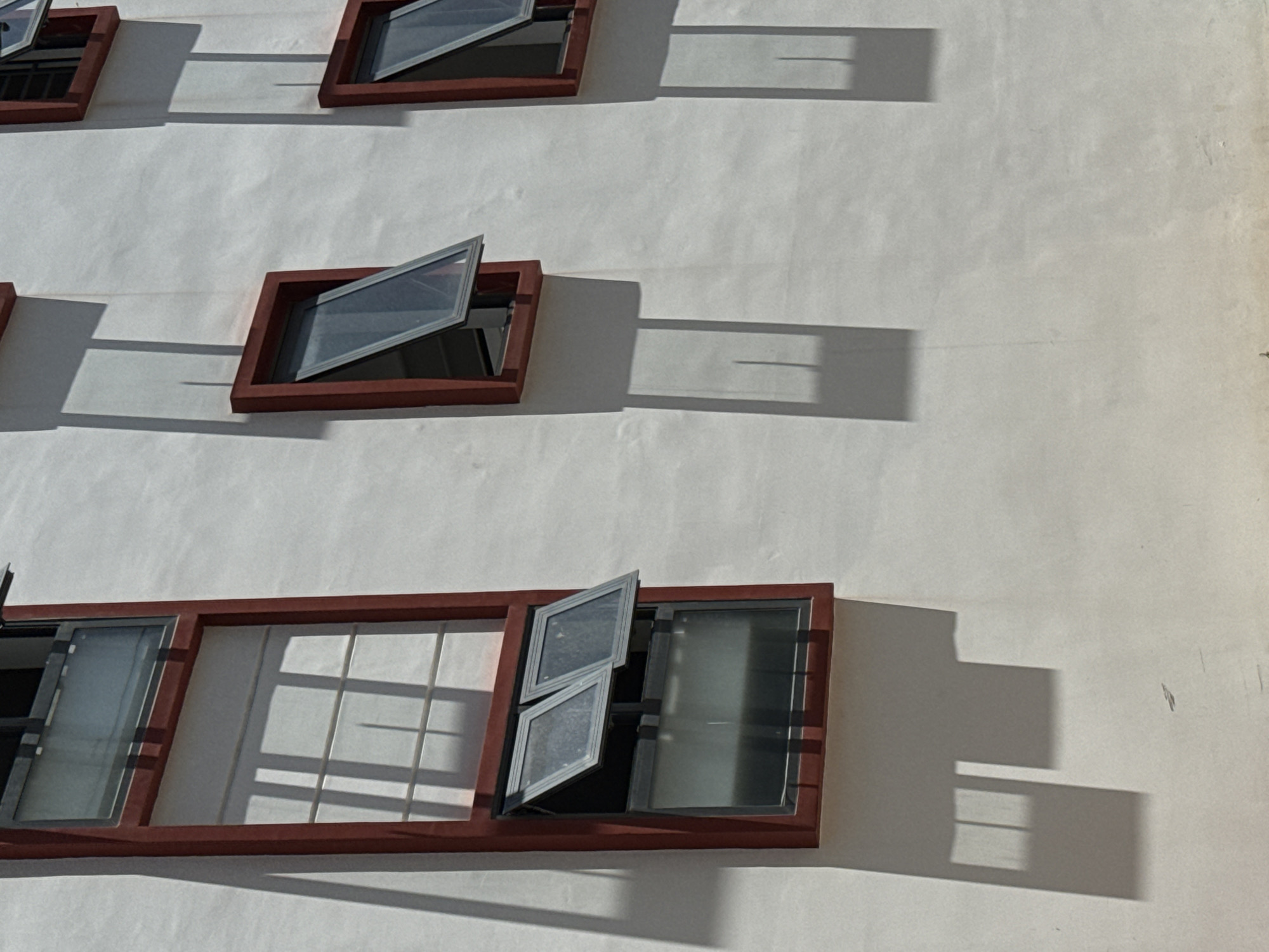 The image displays a section of a white building's exterior wall, featuring three windows with red frames. The windows are of varying sizes and orientations, with some partially open. The sunlight casts distinct shadows of the windows and their frames onto the textured white surface of the wall, creating a pattern of light and dark areas. The scene appears to be an architectural detail within a city setting, however, no specific location in Bac Nha Trang Ward, Vietnam, can be determined from the visual information provided. There are no people, animals, or other significant objects visible in the frame.