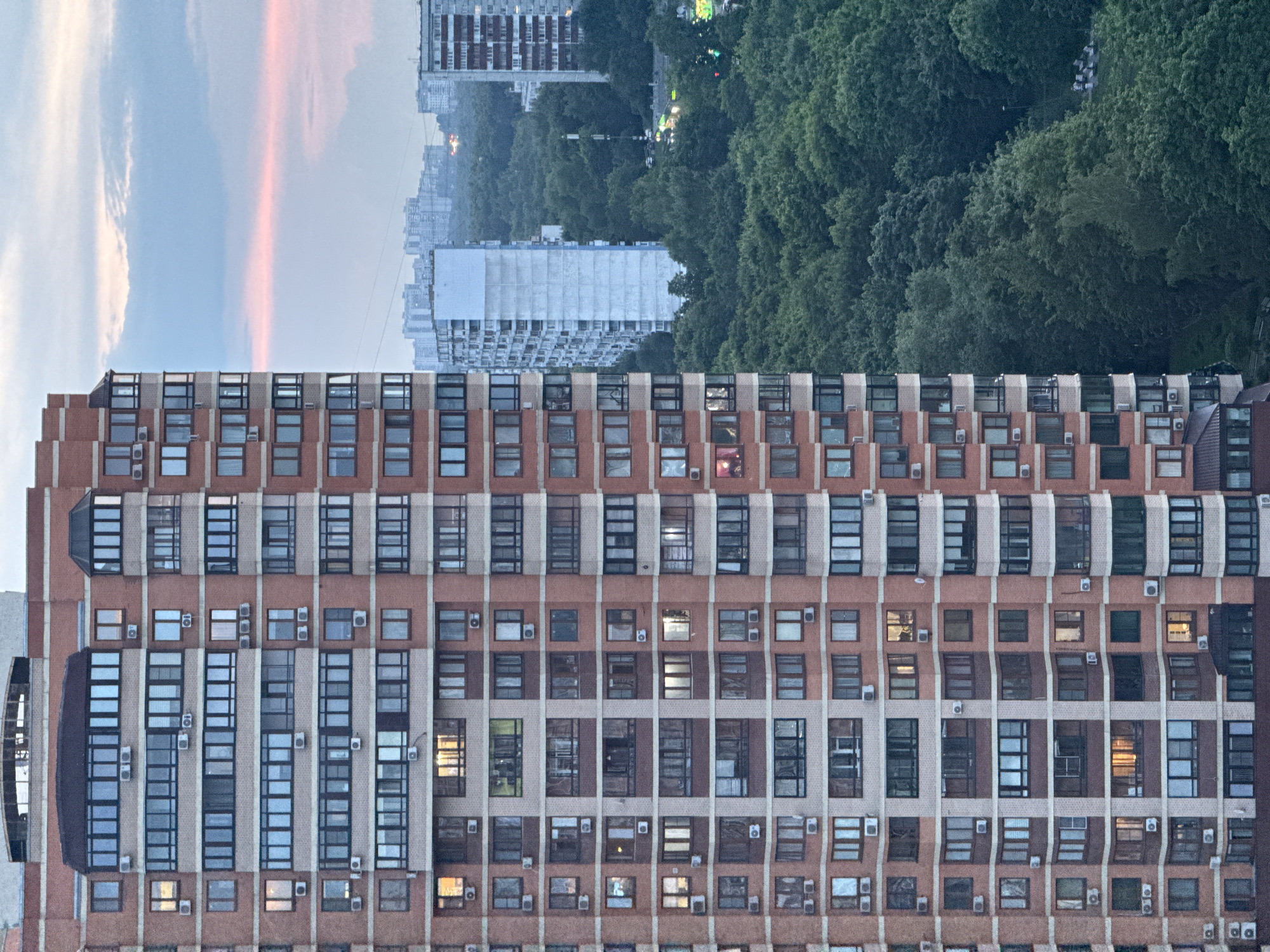 The image shows a tall residential building in Moscow, Russia, on a cloudy day. The building is made of red brick and has many windows and balconies. There are other buildings and trees in the background. The sky is a pale pink and blue. The image is taken from a high angle, looking down at the building and the surrounding area.