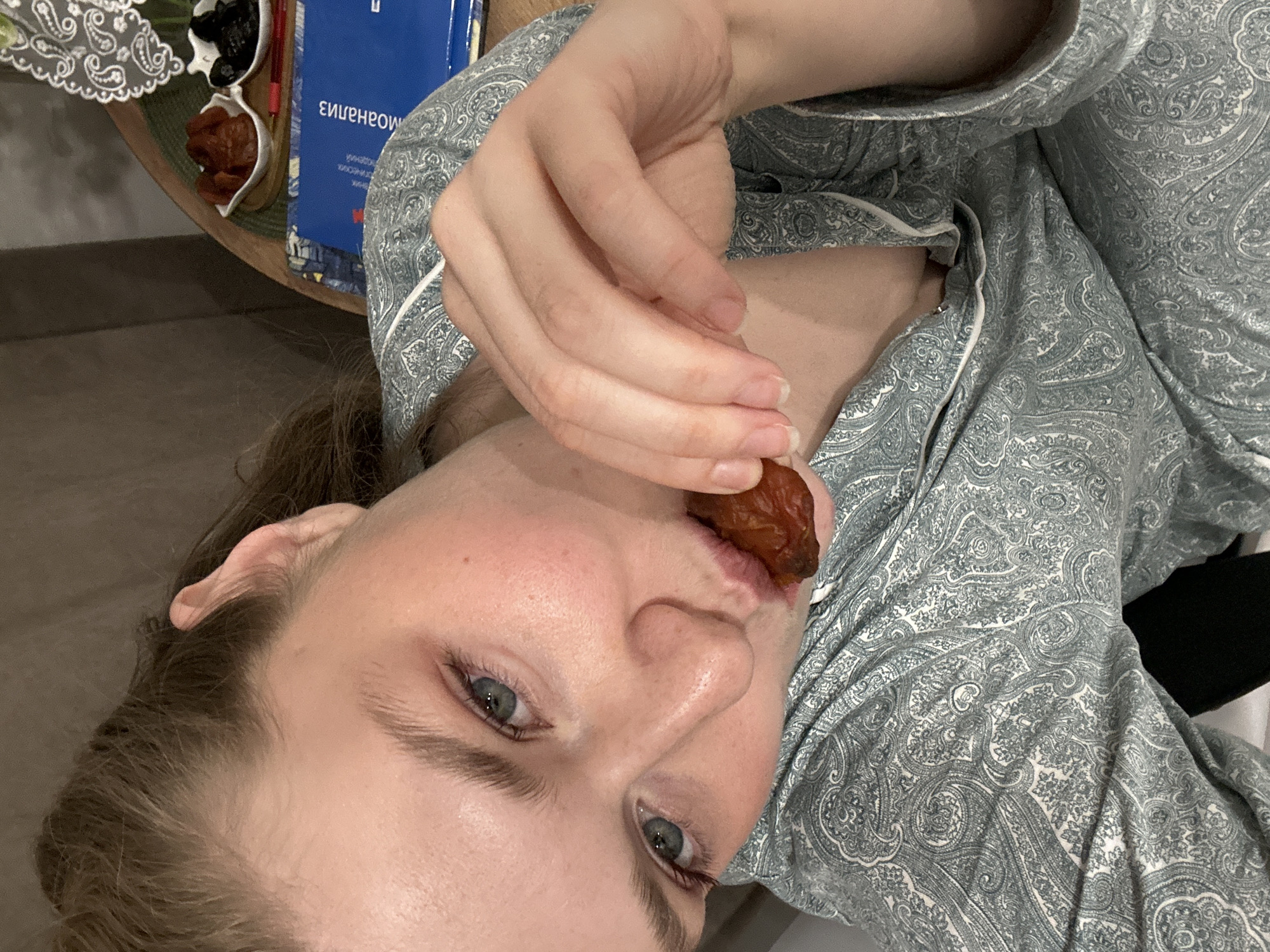 A woman with fair skin and blue eyes is holding a date to her lips, presumably about to eat it. She is wearing a paisley-patterned pajama top with white trim. In the background, on a round table, there is a dish of dried fruit and a blue book with lettering in Russian. The specific location in Kazan, Russia, cannot be determined from this image.