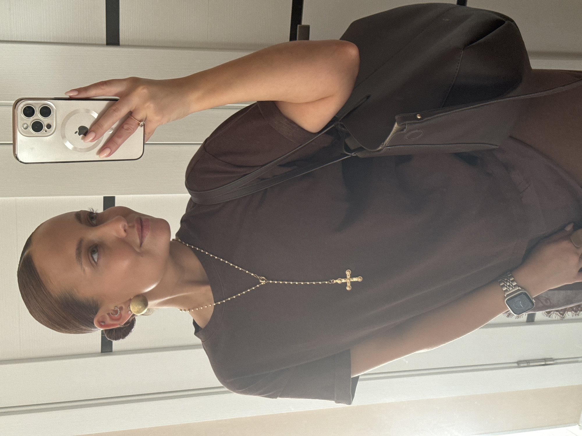 A person is taking a selfie with a smartphone. The individual is wearing a brown t-shirt, a gold cross necklace, a gold wristwatch, and gold earrings. A brown purse hangs over the shoulder. The background appears to be a wall with white vertical panels. While location is difficult, Chaikovsky, Russia is not visually verified.