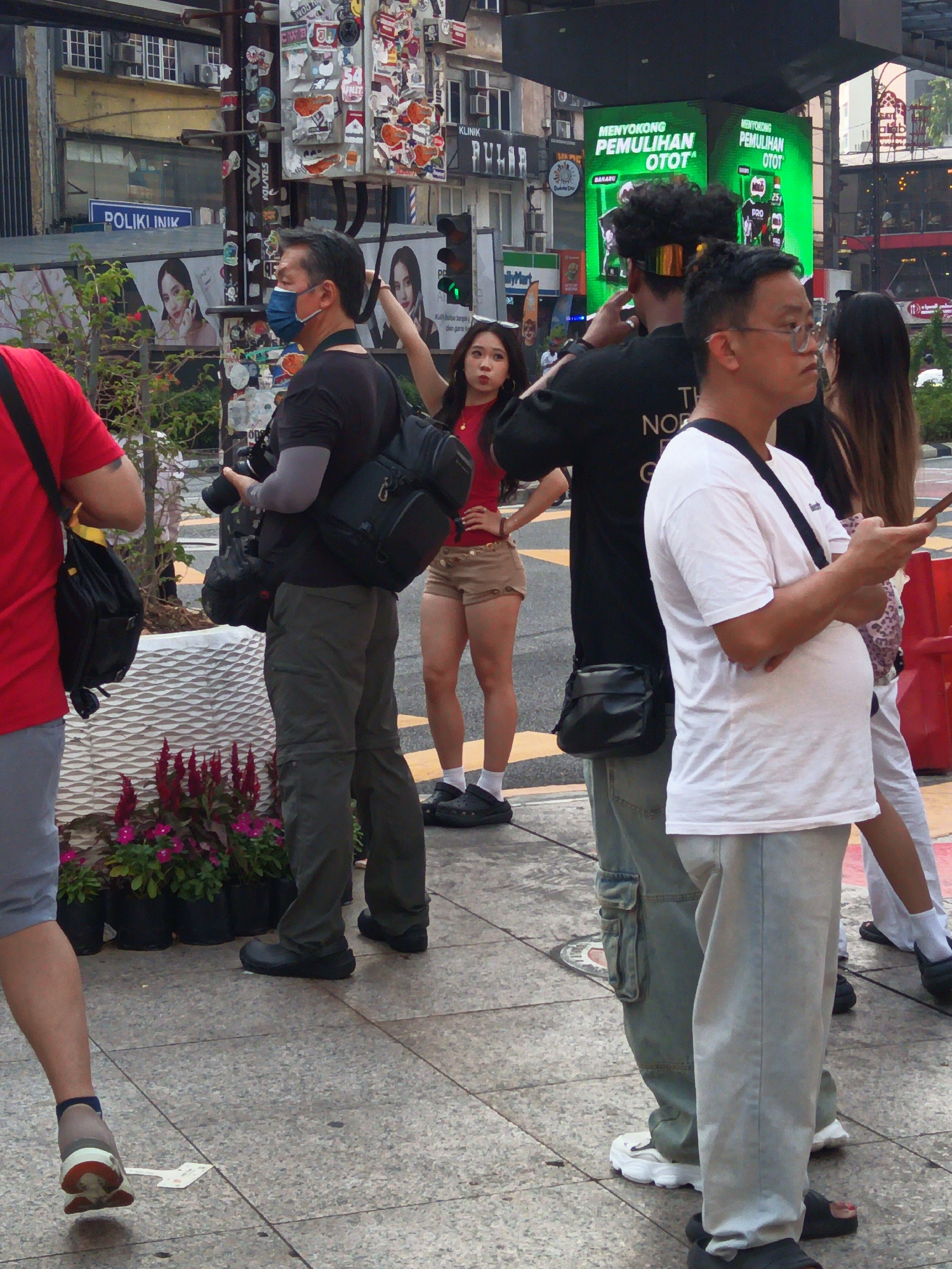The image depicts a street scene in Kuala Lumpur, Malaysia, possibly near Jalan Alor or a similar bustling urban area. In the foreground, a person wearing a red t-shirt and blue shorts is partially visible on the left. To their right, a man in a black t-shirt and gray cargo pants is holding a camera, with a black backpack slung over his shoulder. He appears to be wearing a blue face mask. In the middle ground, a woman in a red top and beige shorts stands with her hands on her hips. Next to her, a person with curly dark hair and a gold headband is looking to the right. Further to the right, a man in a white t-shirt and light blue jeans is looking to the side, holding a smartphone. Behind them, a large green digital billboard displays advertisements in Malay. The environment features a sidewalk with decorative planters containing red and pink flowers. Buildings with windows and air conditioning units are visible in the background. The overall scene suggests a public space with people engaged in various activities, possibly documenting or observing the surroundings.