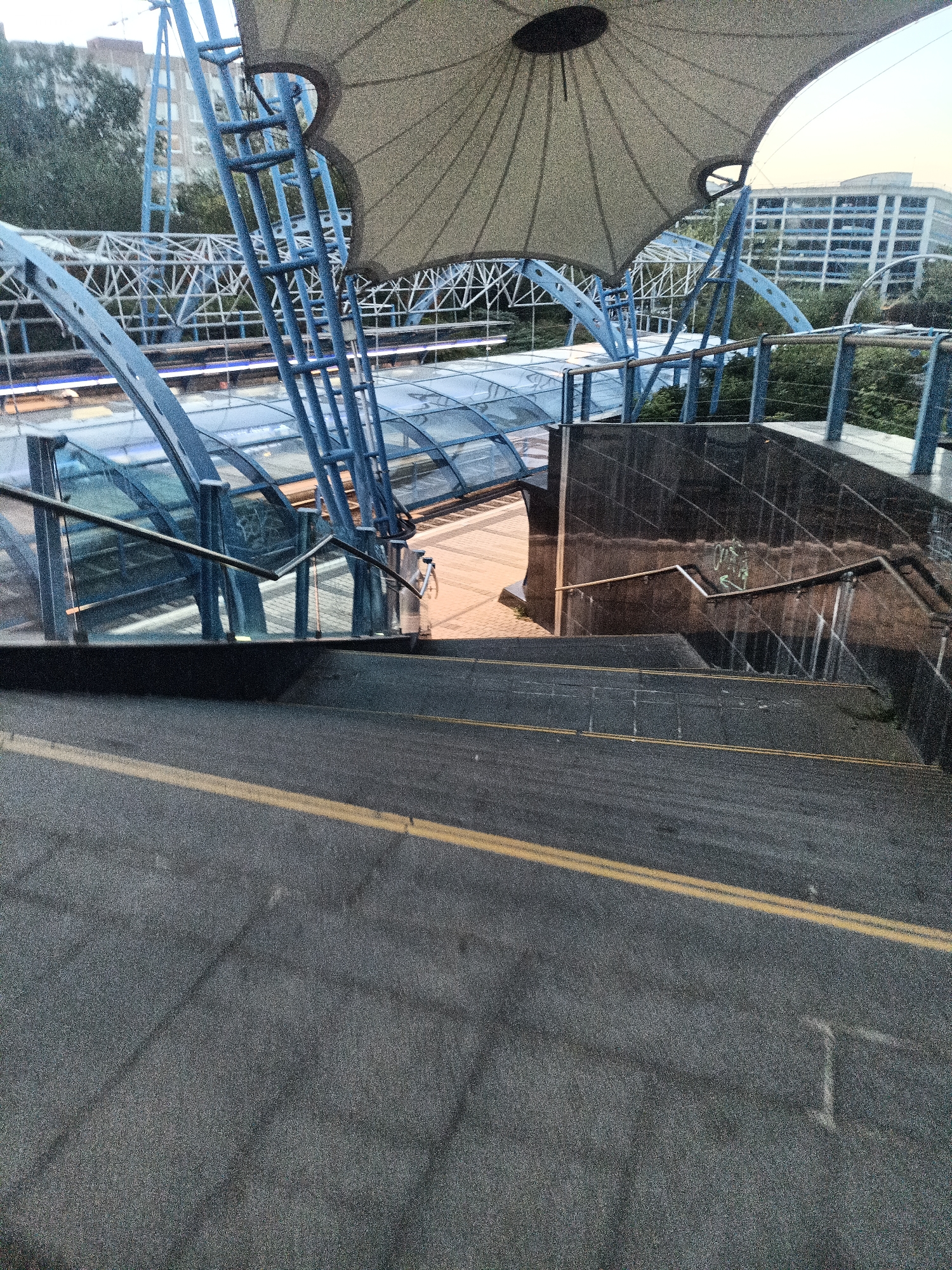 The image shows an outdoor staircase leading down towards a covered walkway and train tracks. The staircase is made of asphalt with two yellow lines painted on the surface. A dark, glossy wall with a diamond pattern is visible to the right of the stairs. Above the walkway, a large, light-colored canopy is supported by blue metal structures. Train tracks and platforms are visible beneath the walkway, which is enclosed by glass and blue metal supports. In the background, trees and a multi-story building with numerous windows can be seen. The sky appears overcast. There are no people visible in the image. This scene is consistent with an outdoor train station or public transport hub. Based on the architectural style and surrounding environment, this location could be a modern train station in a city.