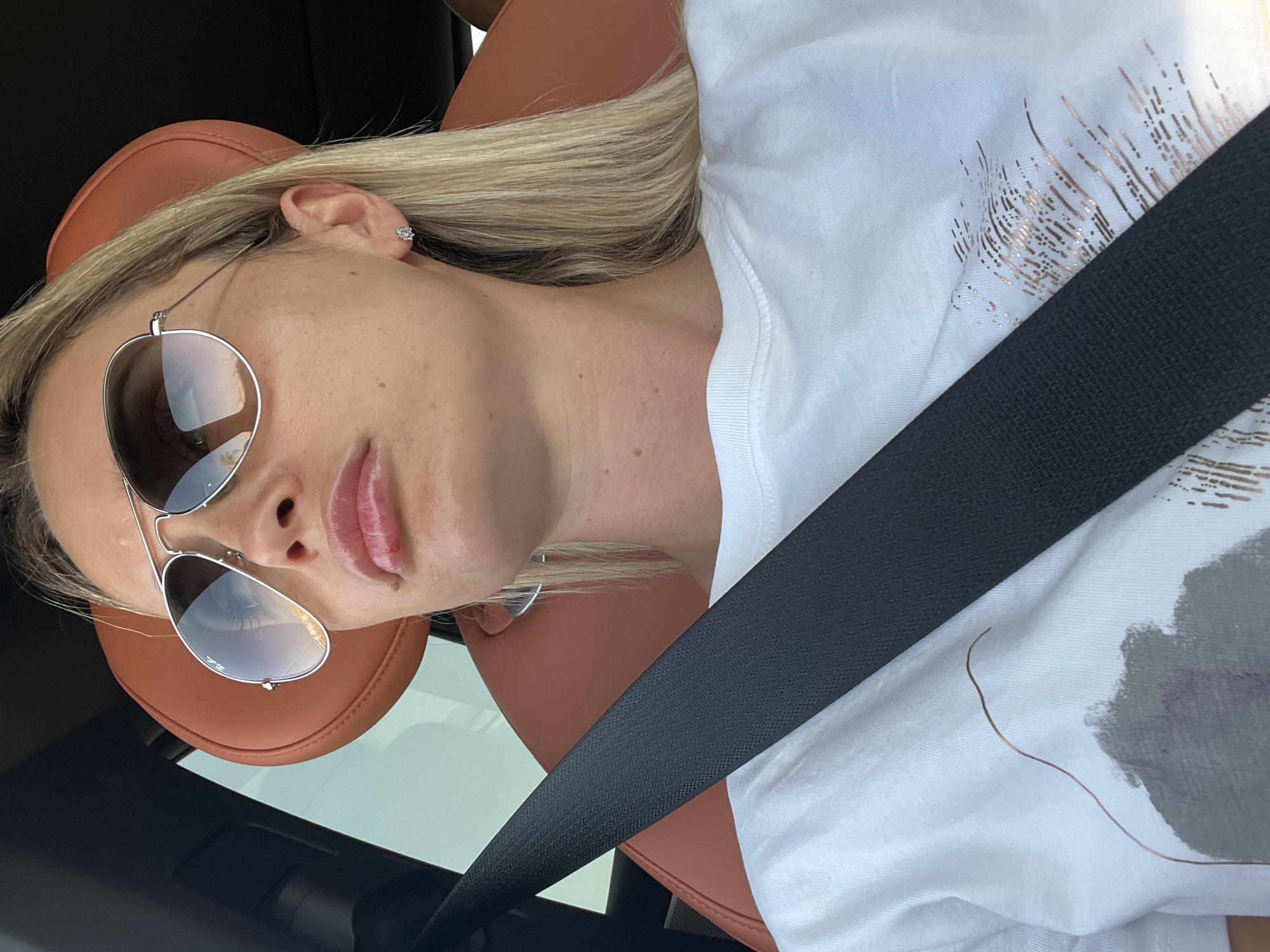In an indoor shot, a woman is seen from the chest up, seated in a vehicle, with a black seatbelt prominently displayed across her body. She wears large, silver-rimmed aviator sunglasses with gradient lenses and a white t-shirt adorned with a subtle abstract design. Her fair skin is visible, and her blonde hair is partially visible. The interior of the vehicle appears to be upholstered in a rich brown leather, suggesting a luxurious setting. The background is mostly dark, with a hint of what looks like a car headrest. The lighting is bright, casting a natural, daytime ambiance. No specific location in Sharm El Sheikh, Egypt, can be determined from the visual information provided.