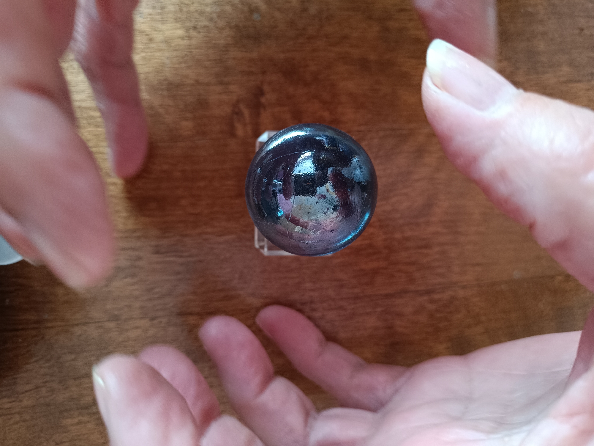 A clear glass sphere, reflecting light and surrounding hands, sits centered on a wooden surface. Four hands are visible, two on the left and two on the right, partially framing the sphere. The sphere appears to be resting on a small, clear stand. The wooden surface has a visible grain pattern. No specific location in Tebras, Latvia is discernible from the image.