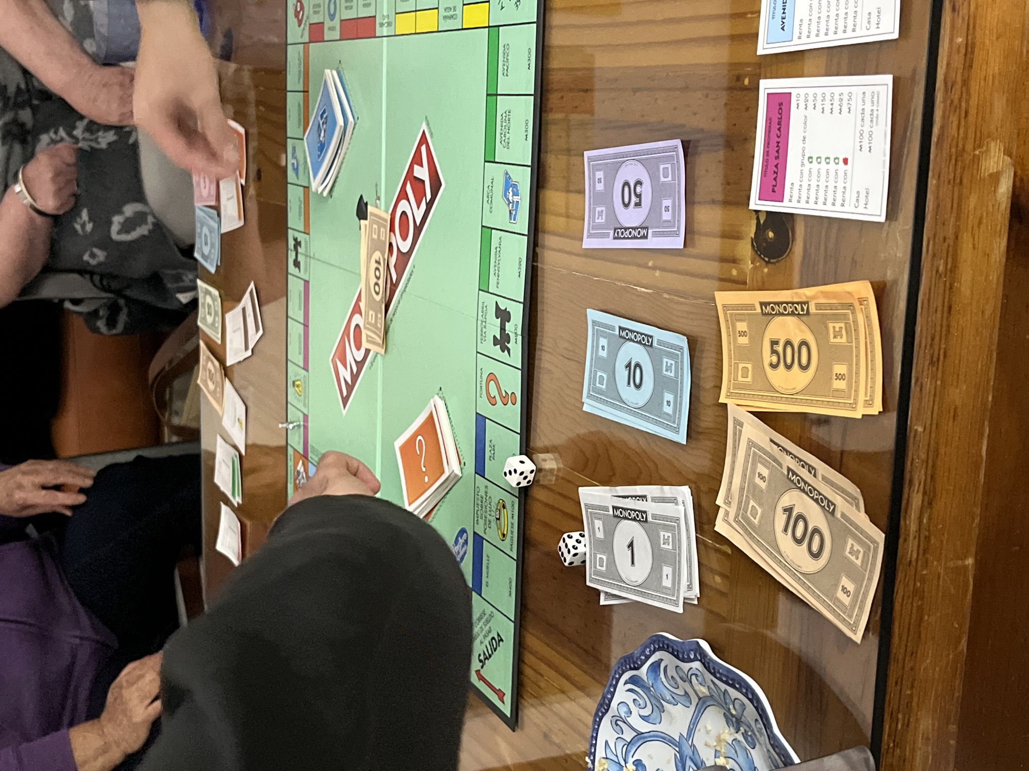 A game of Monopoly is in progress on a reflective wooden table. People are seated around the table, their hands visible as they play. A Monopoly board is at the center, with stacks of property cards arranged around it. Dice are scattered near the "FORTUNA" and "SALIDA" spaces. Monopoly money in denominations of 1, 10, 50, 100, and 500 is piled on the table. One property card, "PLAZA SAN CARLOS," is visible, indicating this is a Spanish-language version of the game. Part of a blue and white patterned bowl or dish is in the lower right corner.