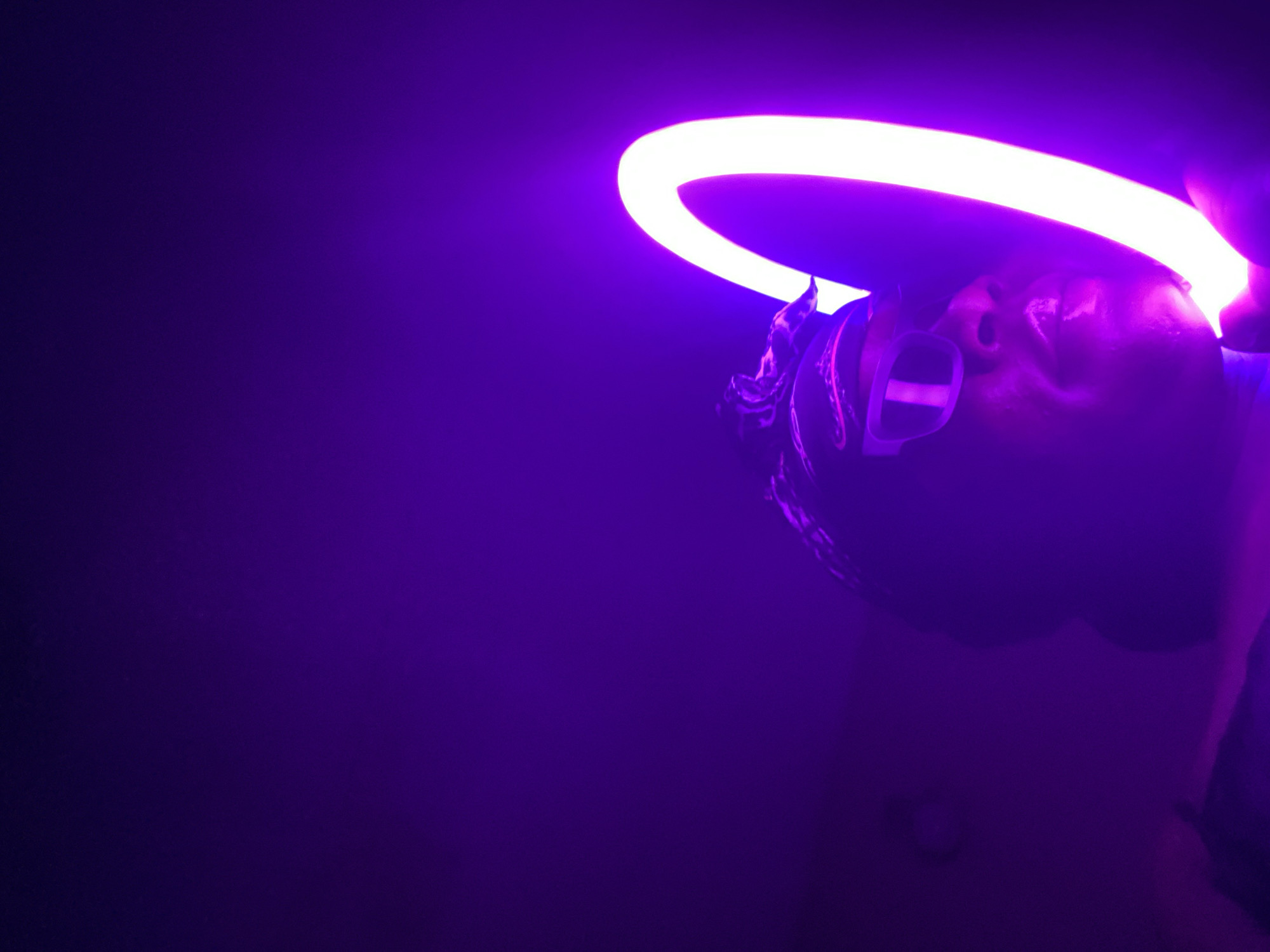This close-up, low-angle shot captures a person illuminated by a vibrant purple ring light in a dark setting, likely indoors in Maigizo, Nigeria. The individual, seen from the nose upwards, is positioned beneath or directly in front of the glowing circular light source, which casts an intense purple hue across the scene.
The person's face is partially obscured by the light, but features like the nose and part of the forehead are visible, strongly lit and reflecting the purple glow. They appear to be wearing a dark head covering, possibly a bandana or durag, with some textured patterns discernible. Additionally, the person is wearing distinct rectangular-framed glasses or goggles, with a prominent horizontal bar across the lenses.
The background is a deep, uniform purple, fading into darker shades in the corners, indicating a lack of other light sources and emphasizing the subject and the ring light. The angle of the shot suggests the person is looking directly up into the light or towards the camera positioned below. The image evokes a stylized, perhaps artistic or experimental, portrait or a moment captured during content creation. No specific activity beyond posing or being illuminated is clearly depicted. There is no visible text in the image. The time of day is not discernible, as the artificial lighting creates its own environment.