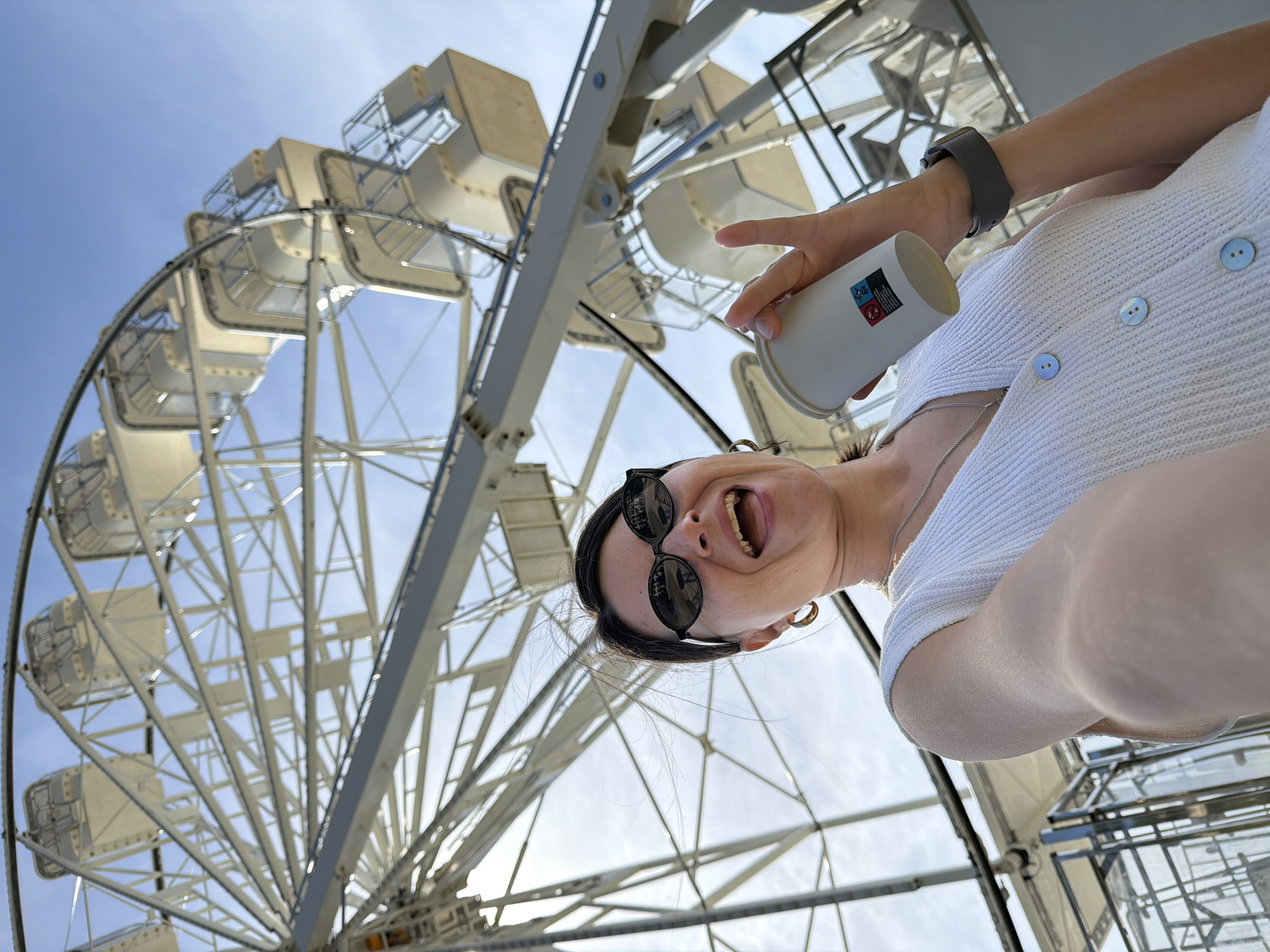 A low-angle shot depicts a woman in Budva, Montenegro, positioned in front of a Ferris wheel on a sunny day. The woman is wearing black sunglasses, a white button-down shirt, and a gray smartwatch. She is holding a white disposable cup in her right hand. Her mouth is open, and she appears to be smiling. The Ferris wheel dominates the background, with a light blue sky visible between its spokes. The location suggests this is likely the Ferris wheel found in Budva.