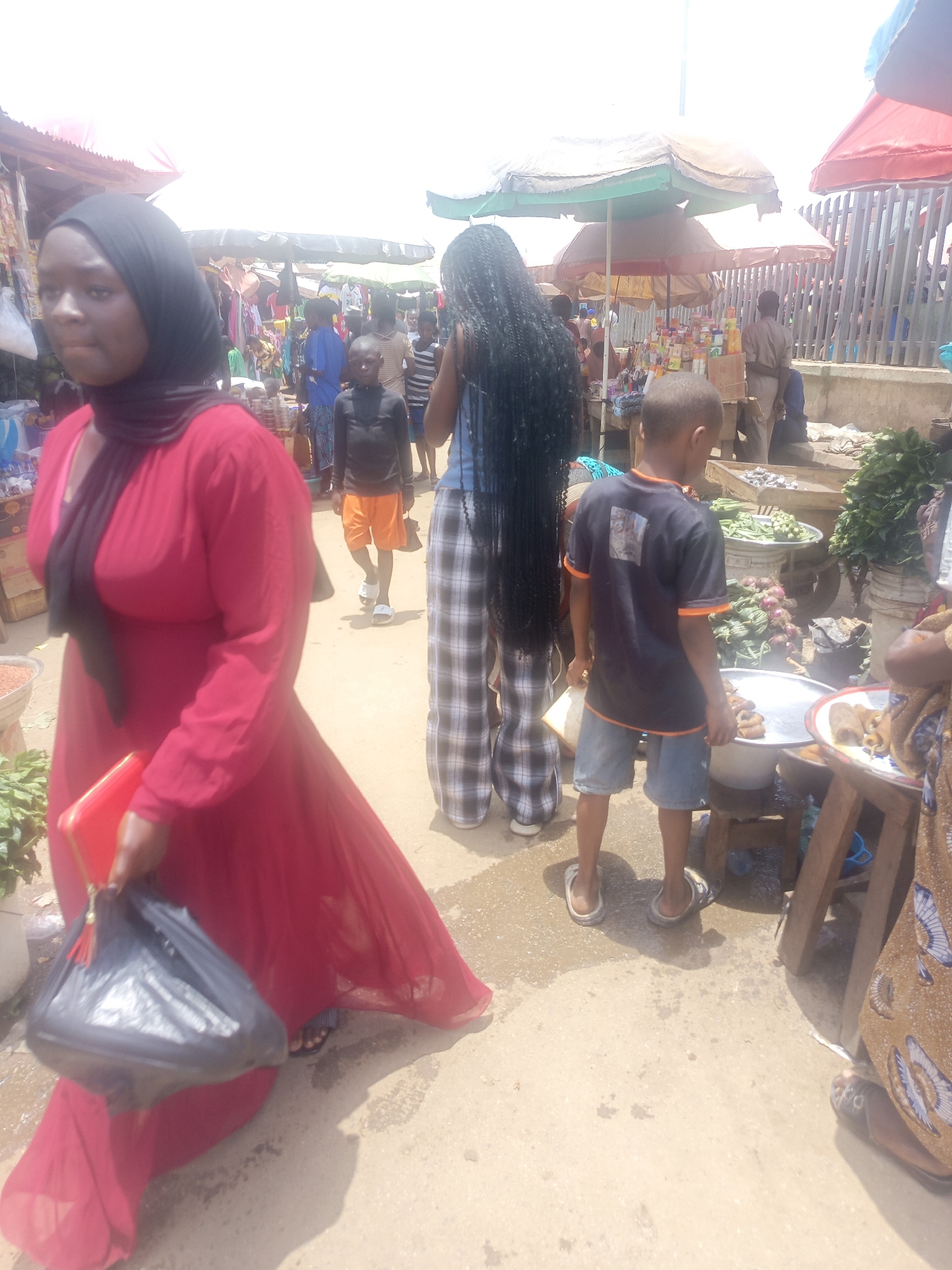 Here's a forensic description of the image:
**Location:** Outdoor market in an urban area. The city is unknown.
**People:**
* An adult female wearing a red dress and headscarf. She is holding a red clutch and a black plastic bag.
* A person with long black hair, wearing a blue top and checkered pants.
* Two young boys are present. One is wearing a black shirt and jean shorts, and the other a black shirt with orange shorts.
* Other individuals are visible in the background.
**Objects:**
* Market stalls with produce and goods.
* Umbrellas shading the stalls.
* Plastic bags, clothing, food items.
* Concrete ground.
**Actions/Interactions:**
* People are walking through the market.
* The individuals are either shopping or working at the market stalls.
**Environment:**
* Bright daylight.
* The market appears to be crowded.
* Ground is paved and wet in some areas.