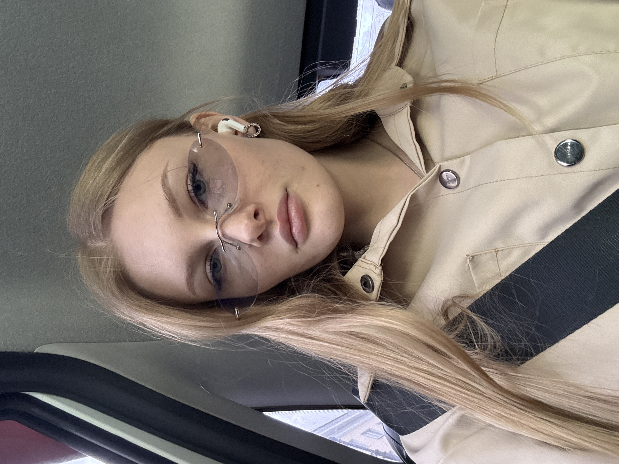 A young woman with blonde hair is wearing rimless glasses and a tan shirt with a black stripe, seated inside a vehicle. She also wears wireless earbuds. The image was taken at an angle, with the vehicle's window and frame visible. Without additional information, the location in Perm, Russia, cannot be confirmed or determined.