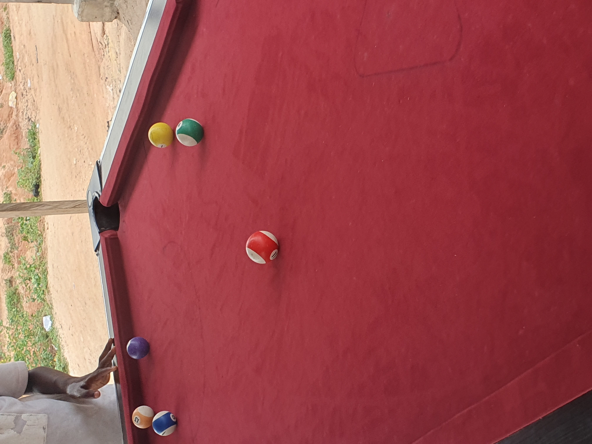 The image shows a close-up, high-angle view of a portion of a red billiard table. Several billiard balls are scattered on the felt. From left to right, there's a yellow ball next to a green ball with white stripes, a red and white striped ball, and a purple ball. Closer to the bottom of the frame are an orange ball with a white number 13 and a blue and white striped ball. A person's arm and hand are visible on the left side of the image, positioned near the purple and orange balls, suggesting they might be about to play a shot. The background is out of focus, showing a sandy, dirt-like ground with some sparse green vegetation, and a concrete structure that could be part of a building or support. The lighting suggests it is daytime, with soft, natural light illuminating the scene. The setting appears to be outdoors or in an open-air covered area. There is no visible text in the image.