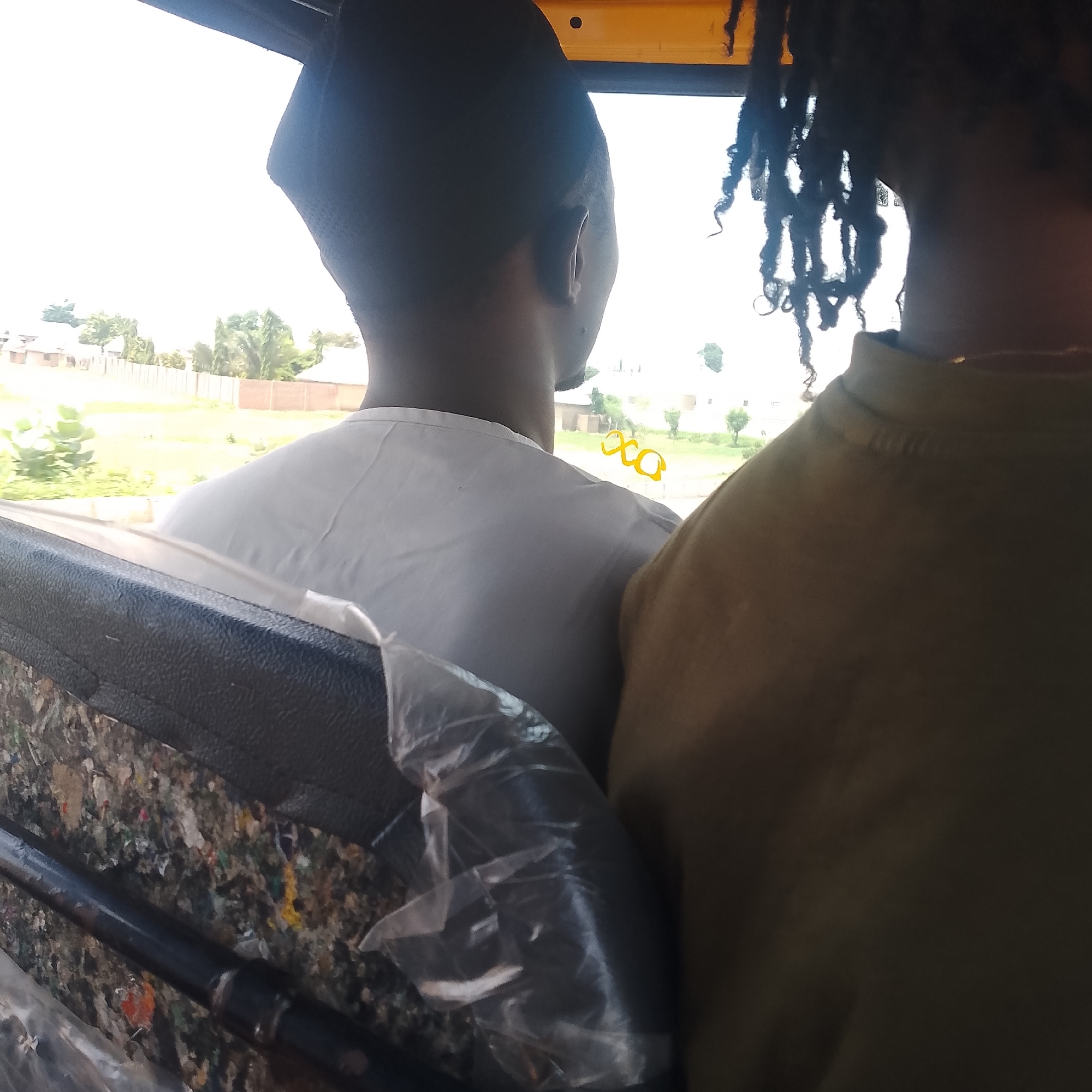This image shows the backs of two individuals sitting in what appears to be the passenger area of a vehicle, possibly a bus or a tuk-tuk, with an open window offering a view of the outside. The person in the foreground is wearing a white t-shirt and has short hair. To their right, a second person is visible, wearing a khaki-colored shirt and sporting dreadlocks.
The scene outside the window suggests a daytime setting, with bright, possibly overcast skies. The landscape features buildings that appear to be houses or small structures, a fence, and vegetation including trees. The overall impression is of a rural or semi-urban environment in Jalingo, Nigeria.
A notable detail is the presence of a black, textured object in the lower part of the image, partially covered by a clear plastic sheet. This object seems to be part of the vehicle's interior, possibly a seat or a storage area, with visible debris or colorful material beneath the plastic. Yellow lettering, resembling "XCO" or similar, is visible on the window behind the person in the white t-shirt. No specific emotions are discernible as the subjects are seen from behind.