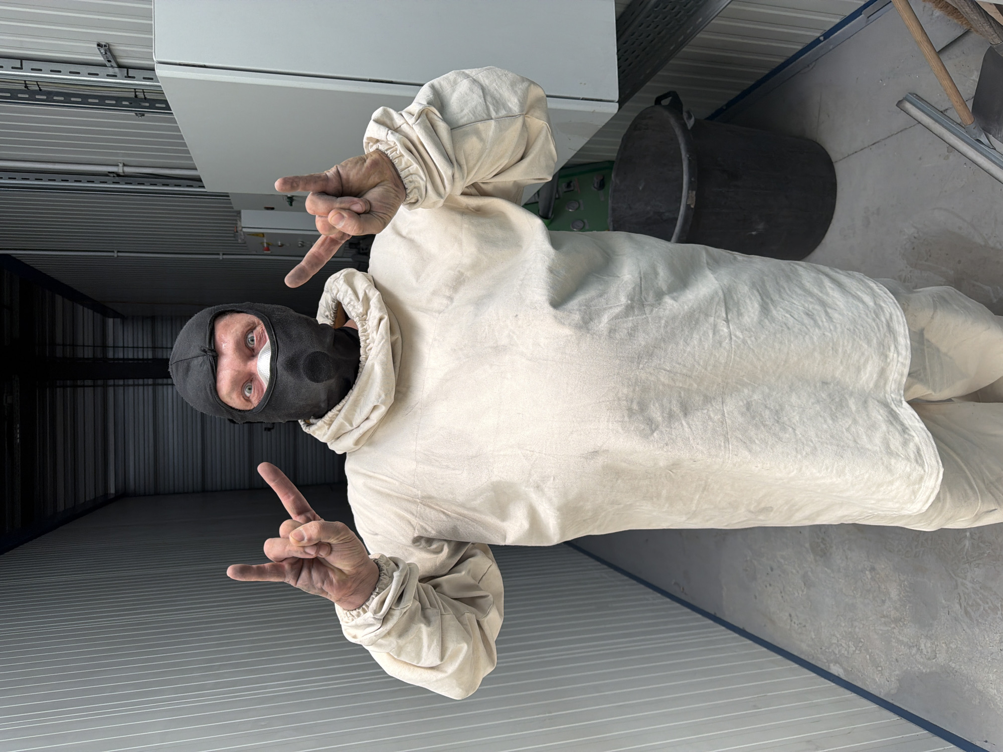 A person, wearing a tan protective suit and a black balaclava with a mask, stands in what appears to be a storage unit or industrial space in Mol, Belgium. The person is making a "rock on" hand gesture with both hands, with their thumbs extended and index and pinky fingers raised. Their eyes are visible through the mask and appear wide open, looking directly at the camera. The suit is loose-fitting and appears to be made of a textured fabric. The background consists of light-colored corrugated metal walls, a concrete floor, and some industrial equipment, including a large black plastic bin and what looks like electrical panels. The lighting is somewhat dim, suggesting an indoor environment. The overall impression is of a person in protective gear making a defiant or celebratory gesture in a utilitarian setting.