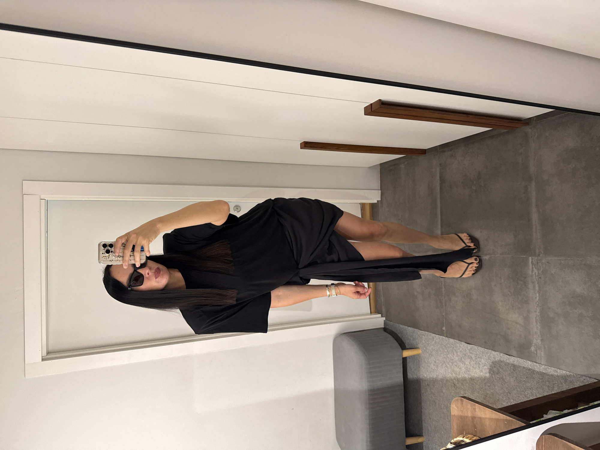 A woman with long black hair and sunglasses is taking a mirror selfie. She is wearing a black t-shirt dress with a tie waist and black thong sandals. The background appears to be a fitting room or dressing area, with a gray bench, a white door, and a full-length mirror. There is no indication of the city of Chaikovsky, Russia in the image.