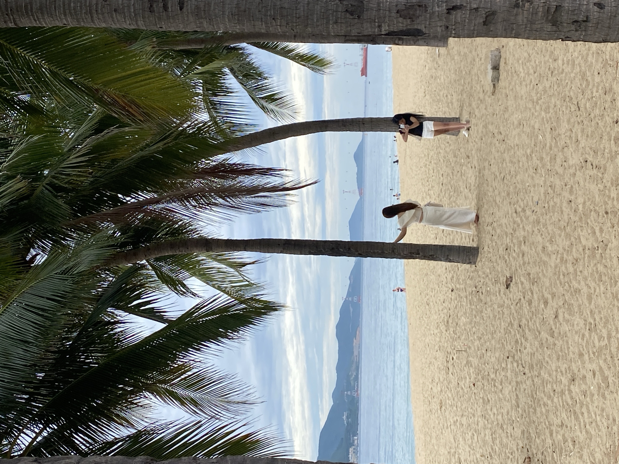 This image depicts a serene beach scene in Nha Trang, Vietnam, likely located along Tran Phu Beach, known for its palm-lined promenade. In the foreground, two women are visible on the sandy shore, framed by the trunks and fronds of mature palm trees. The woman on the left, dressed in an elegant white outfit, stands facing away from the camera, her posture suggesting she might be posing or taking a photo. To her right, a younger woman in a black top and white shorts is actively taking a picture with her phone.
Beyond the immediate foreground, the tranquil blue waters of the sea stretch towards a distant coastline characterized by rolling hills and mountains. The sky is partly cloudy, with a blend of white and gray clouds, suggesting it might be a mild day. A large red cargo ship is visible on the horizon, adding a touch of industrial presence to the otherwise natural landscape. Several smaller boats and distant figures dot the water and beach, indicating a moderate level of activity. The sand itself appears finely granulated, typical of a well-maintained beach. The overall impression is one of a relaxed tropical vacation setting, with the iconic palm trees framing the view and the ocean providing a calming backdrop.