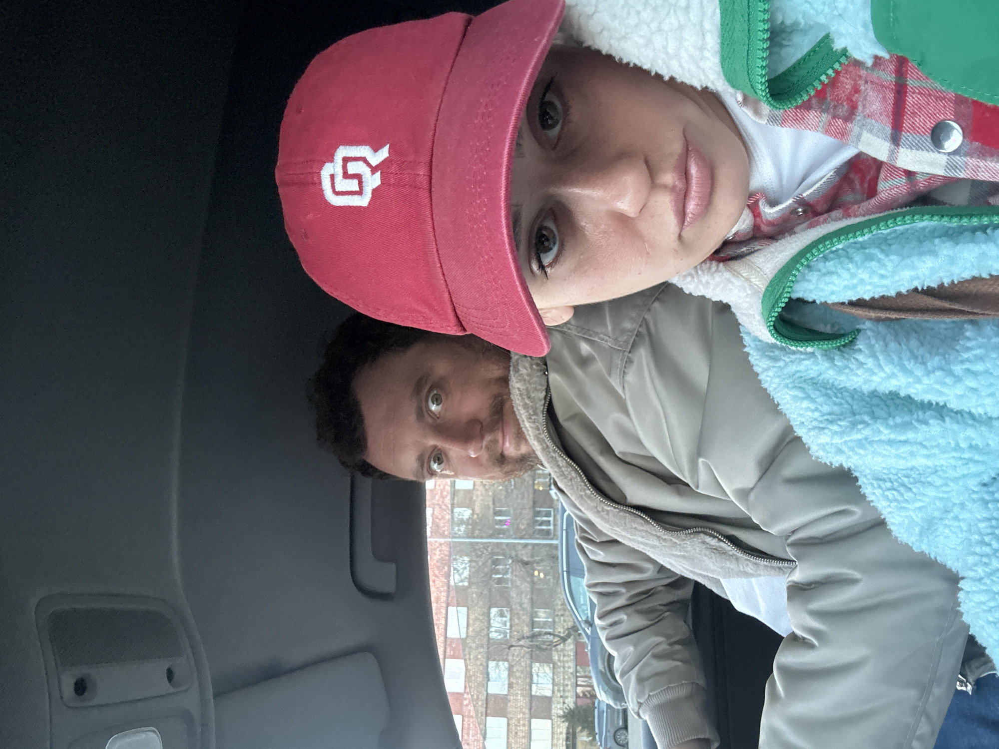Two people are visible inside a vehicle. The individual in the foreground, closer to the camera, is wearing a pink baseball cap with a white "OU" logo. They have light skin and appear to be wearing a light blue fluffy jacket with green trim over a red plaid shirt. The person behind them has dark hair and is wearing a light brown jacket with a white fleece collar. Outside the car window is a multi-story brick building, possibly an apartment building, and a parking area with other vehicles. The location is possibly Krasnoyarsk, Russia. The interior of the car is visible, including parts of the dashboard.