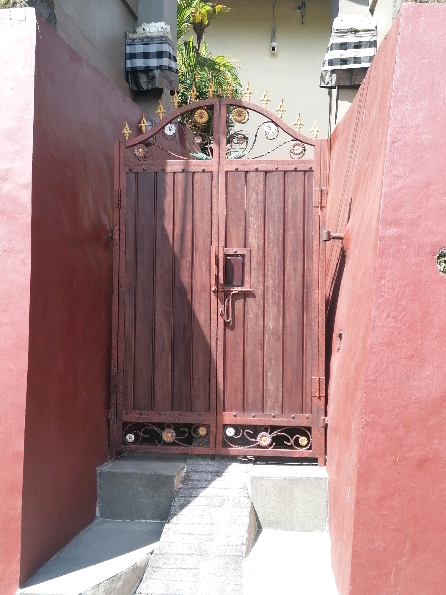 An exterior view of a weathered, reddish-brown double metal gate with vertical wooden planks is captured in the bright midday sun in Ubud District, Indonesia. The gate is adorned with decorative gold metalwork featuring floral motifs and pointed finials at the top. A visible latch mechanism is present on the right side of the left gate. The gate is set within two tall, textured reddish-pink walls, creating a narrow passageway. In front of the gate, there are two concrete steps leading up to it, with a textured concrete ramp or walkway extending outwards. The area above and behind the gate shows parts of a building with a white wall, and a glimpse of greenery and foliage suggests a tropical environment. A security camera is mounted on the upper part of the white wall. The scene is characterized by strong shadows and highlights, indicating direct sunlight.