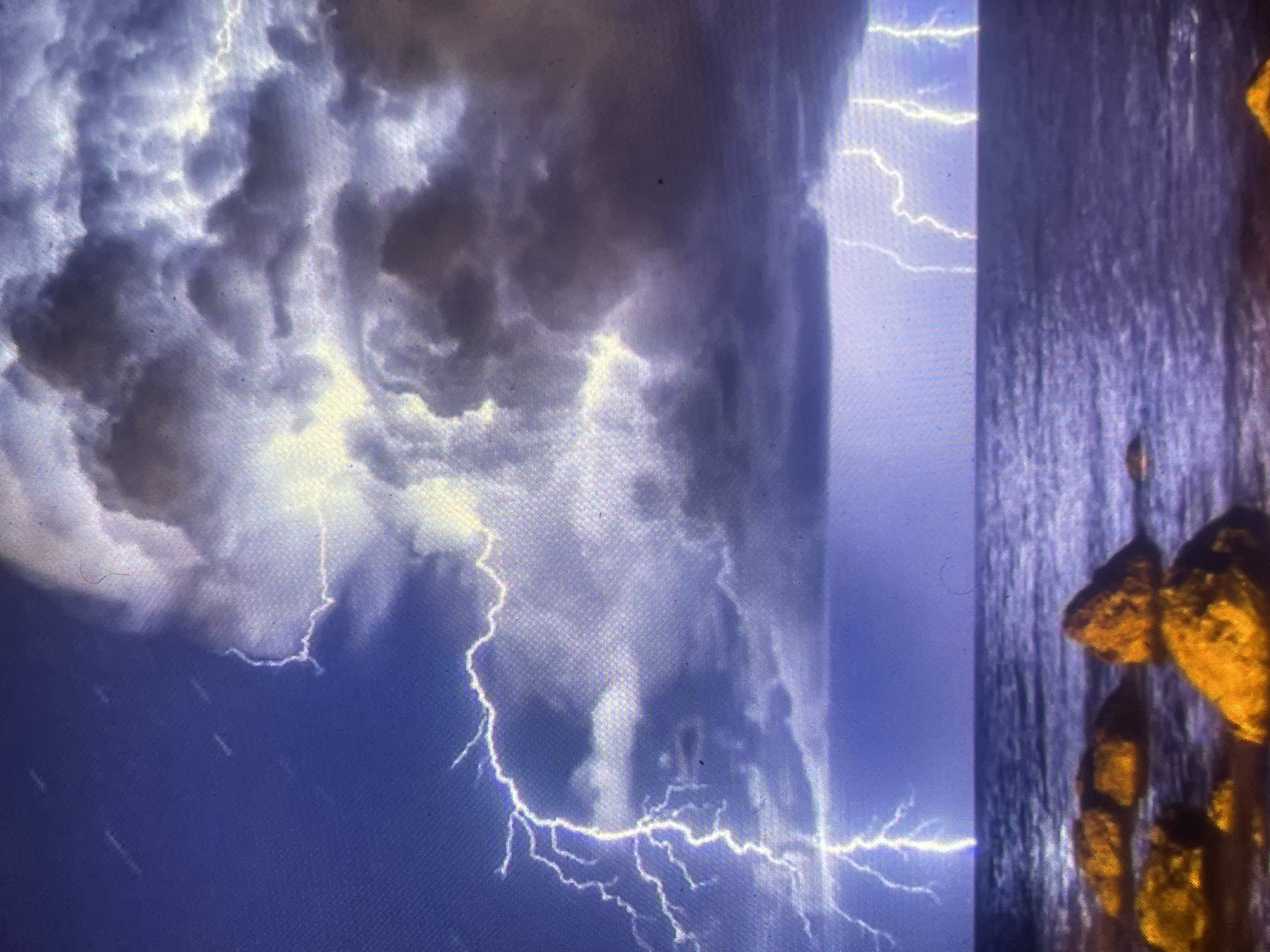 The image depicts a dramatic nighttime or twilight coastal storm scene, identified as being in the vicinity of Zelenyi Hai, Ukraine. The upper sky is dominated by a large mass of turbulent, dark grey and purple-hued clouds, internally illuminated by several bright white and yellow lightning flashes. A prominent, branching lightning bolt descends from the clouds on the left side, appearing to strike near or into the dark blue-purple ocean below. Additional vertical lightning strikes are visible on the far right, illuminating the distant horizon line where the sea meets the stormy sky.
The middle ground is occupied by the dark, choppy surface of the ocean. In the immediate foreground, a scattering of large, rounded rocks, ranging in color from warm orange-brown to darker brown, are partially submerged in the water. The overall lighting is intense but localized to the lightning flashes, creating high contrast between the brightly lit clouds and the darker sea and rocks. The scene is devoid of human subjects, animals, or artificial structures. The image exhibits a visible grid-like pixelation and faint circular artifacts, suggesting it may be a photograph of a screen or a low-resolution digital image.