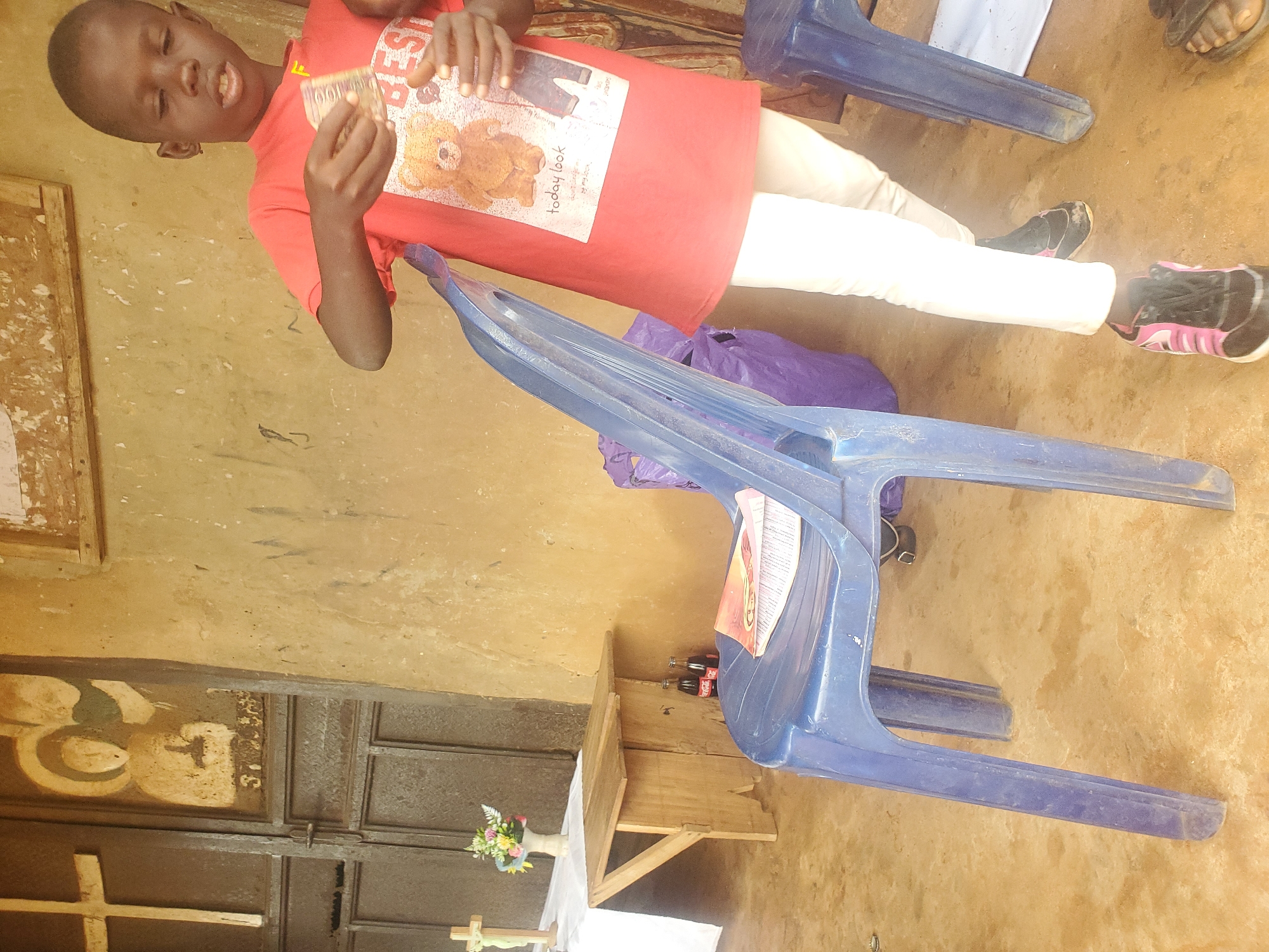 In Nsugpe, Nigeria, a young boy stands in a sparsely decorated room with a dirt floor and plain, earthen-colored walls. He is wearing a red t-shirt with a teddy bear graphic and the words "today look" printed on it, along with white pants and black and pink athletic shoes. In his right hand, he holds a wad of currency. To his left, a blue plastic chair is placed in front of a wooden table. On the chair lies an open book with a colorful cover. Two bottles of Coca-Cola are visible beneath the table. In the background, a metal door is adorned with a cross and a religious painting. The room appears to be part of a place of worship or a community center.