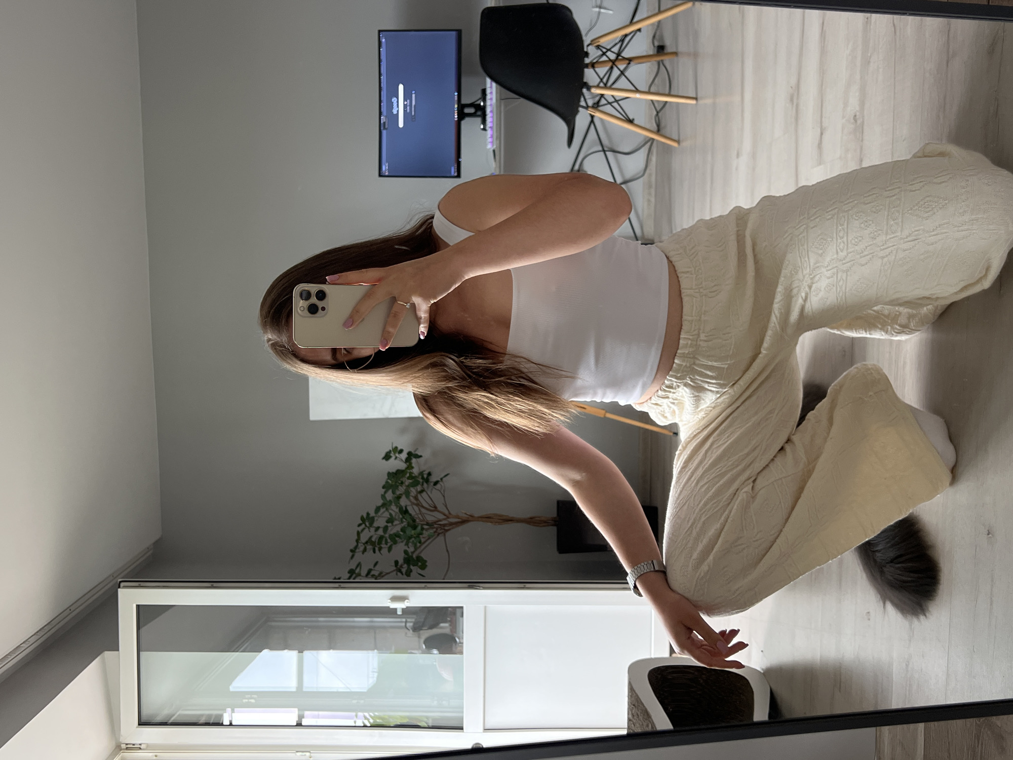 A young woman with long brown hair is crouching in a room, taking a mirror selfie with her phone. She is wearing a white tank top and cream-colored patterned pants. Her left hand is resting on her knee, and her right hand is holding the phone. A gray cat's tail is visible on the floor near her. In the background, there is a computer monitor on a desk, a black chair, a potted plant, and a large glass door that opens to the outside. The room has grey walls and a light-colored floor. The image was taken in Ternopil, Ukraine.