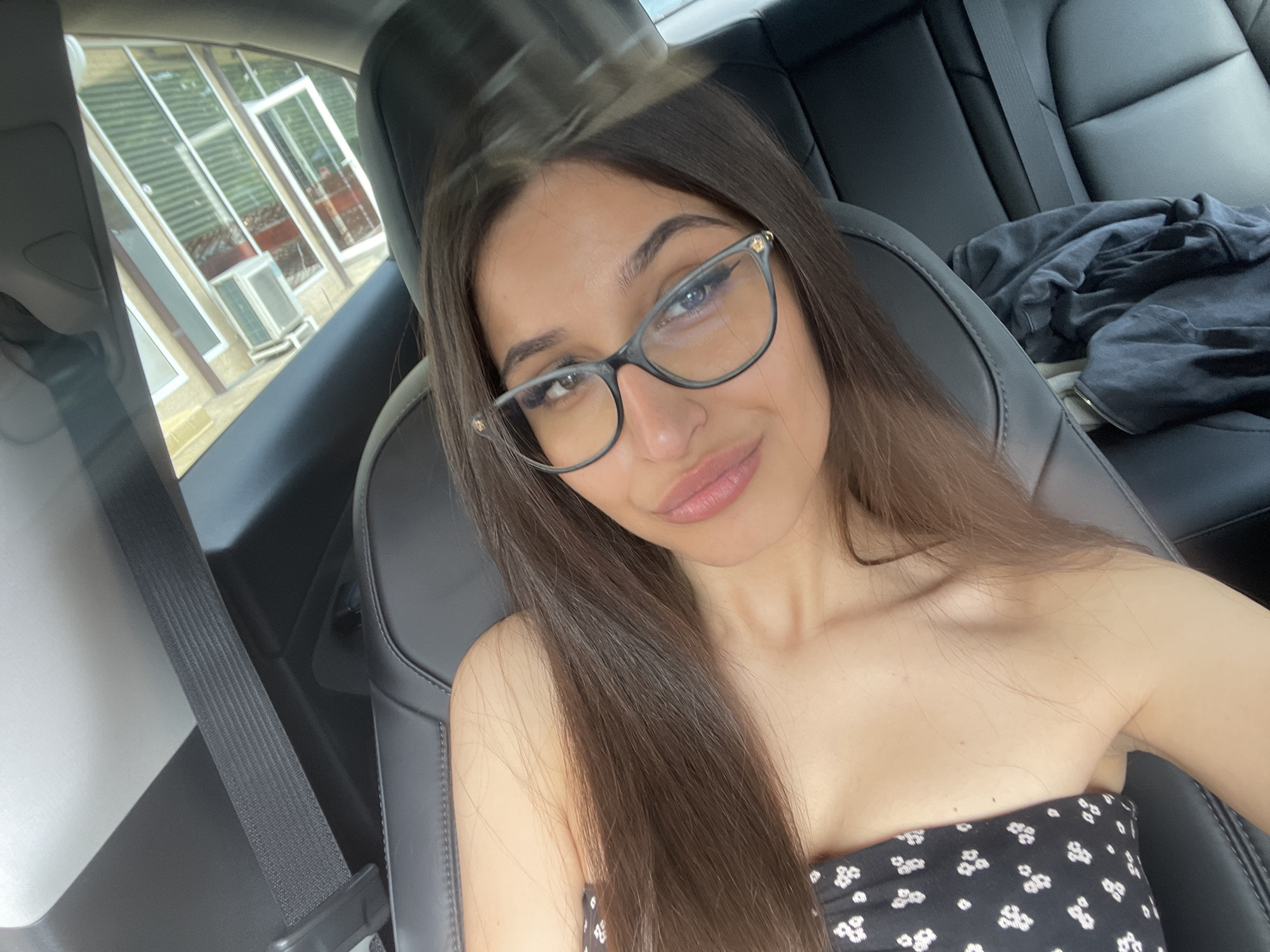 In an interior shot of a car, a young woman with long, dark brown hair and glasses is taking a selfie. She is wearing a black strapless top with a white floral pattern. The car's black leather seats and seatbelt are visible. Through the car window, a building with large windows and an air conditioning unit can be seen. The scene is lit by natural light.