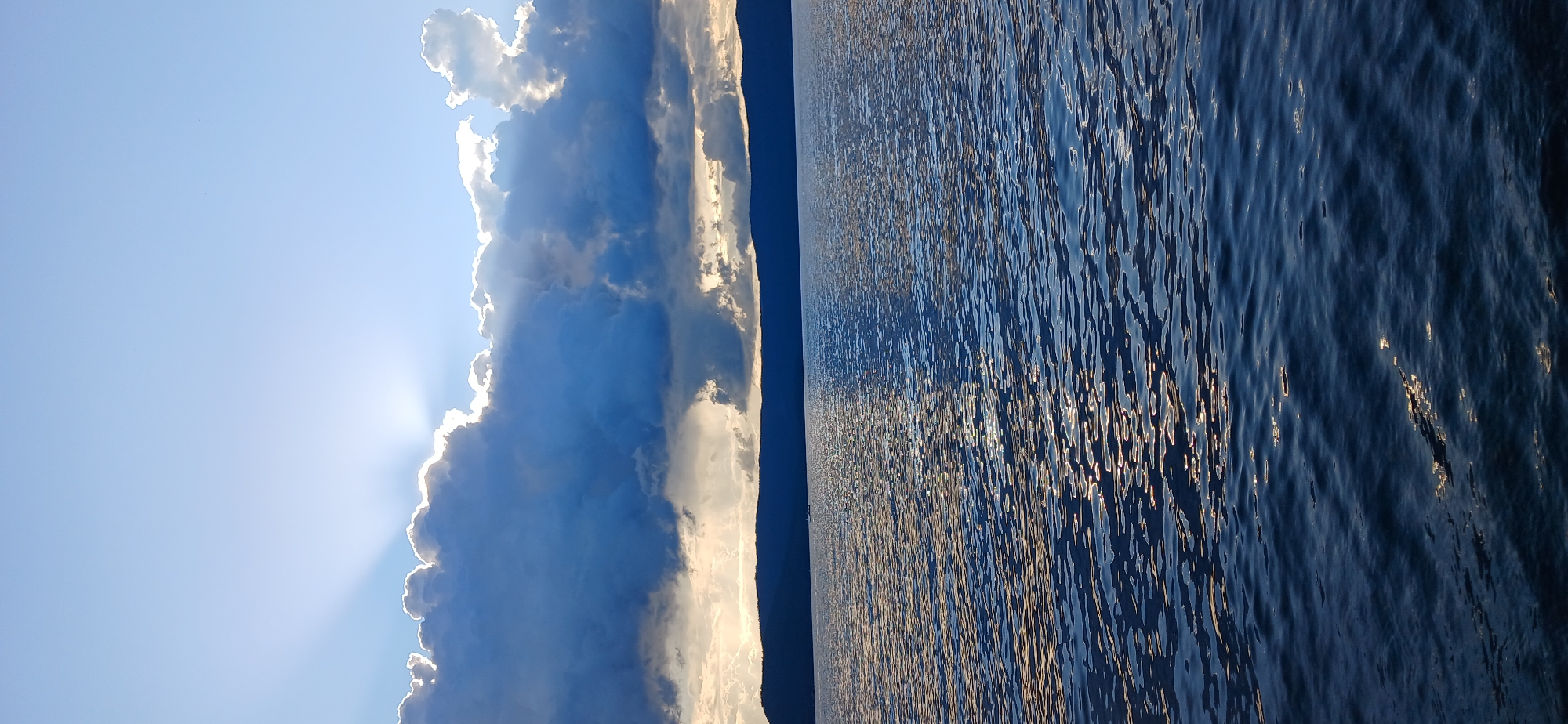 The image depicts a wide shot of a large body of water, likely Lake Turgoyak, in Russia, under a dramatic sky. The water surface is textured with ripples, reflecting the light of the sun peeking through dark, dramatic clouds. These clouds are illuminated from behind, with rays of light fanning out into the bright blue sky. In the background, a dark, silhouetted mountain range runs horizontally across the horizon, separating the water from the sky. The overall impression is one of a serene but powerful natural landscape, possibly at dusk or dawn.