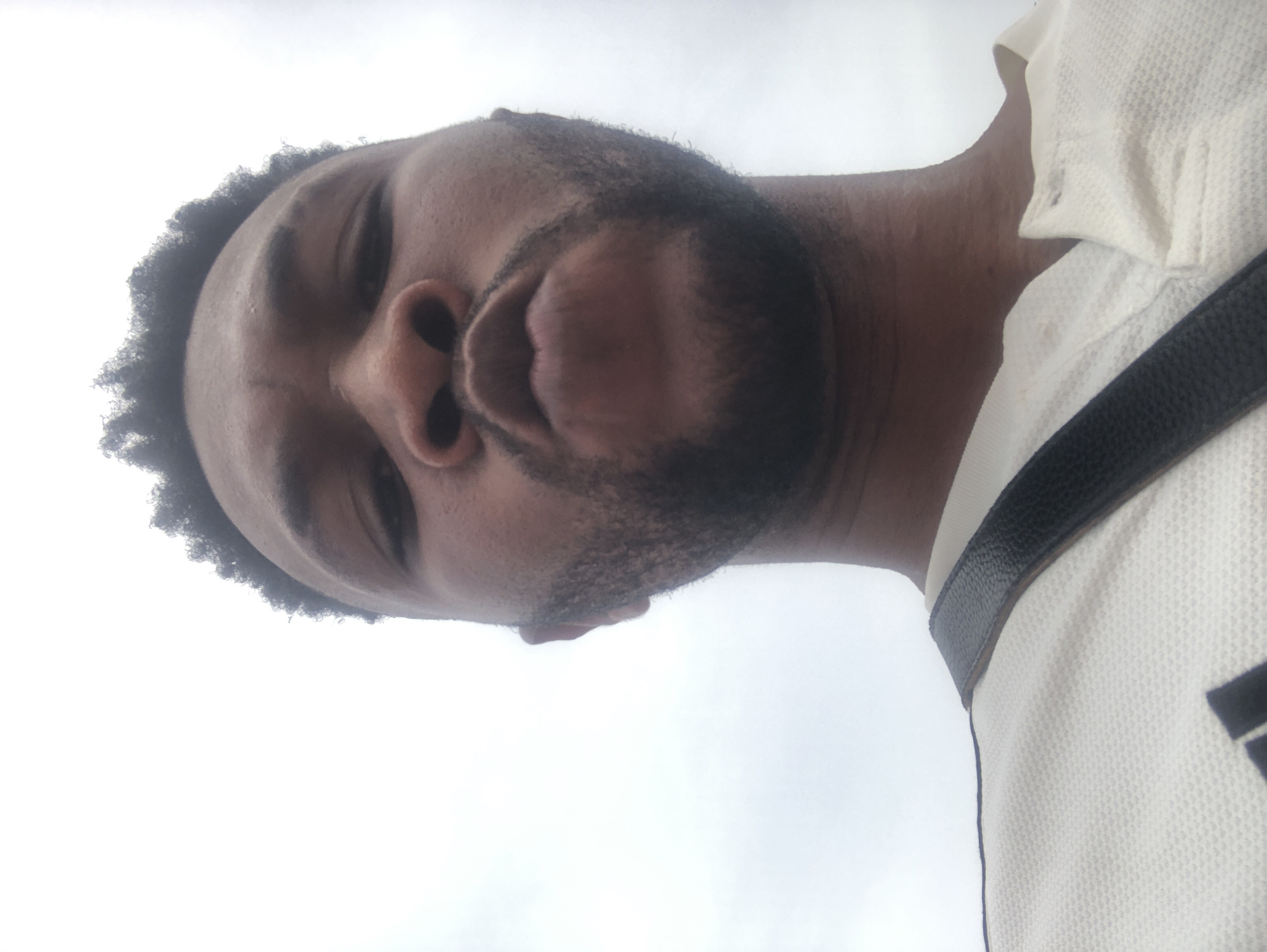 A close-up, upward-facing perspective photograph shows a man with dark skin, a short afro, and a beard. He is wearing a light-colored, collared shirt with a black strap across his chest. The background is a cloudy, overcast sky. The location is not discernible from the image.