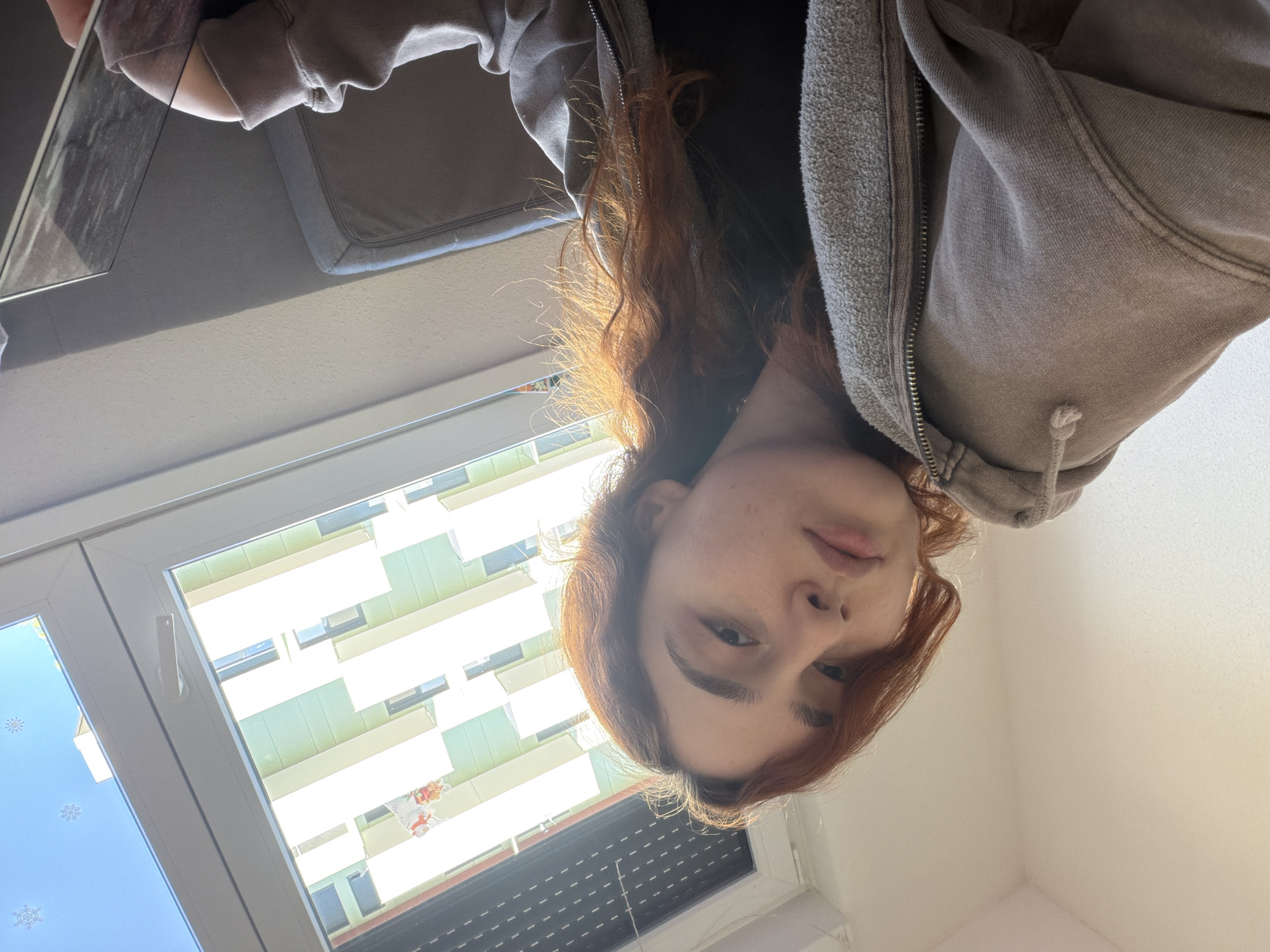 A woman with red hair wearing a gray hooded sweatshirt is looking down at the camera. She is positioned near a window with buildings visible outside, likely in Strasbourg, France given the architectural style. The window is decorated with snowflake stickers. She appears to be holding a tablet or similar device.