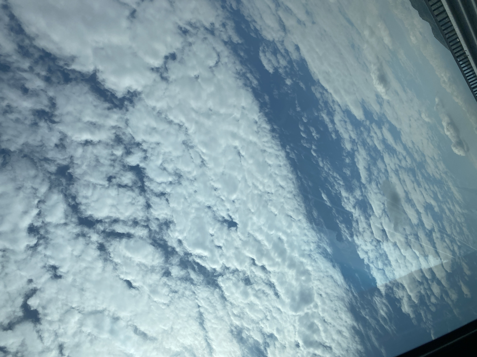 The image shows a sky filled with altocumulus clouds. Patches of blue sky are visible between the cloud formations. The scene is viewed through a window, as indicated by a reflection at the lower right corner and a section of an unidentified structure at the upper right. There is no indication that the image was taken in Sandanski, Bulgaria, or any city for that matter, and there are no people visible.