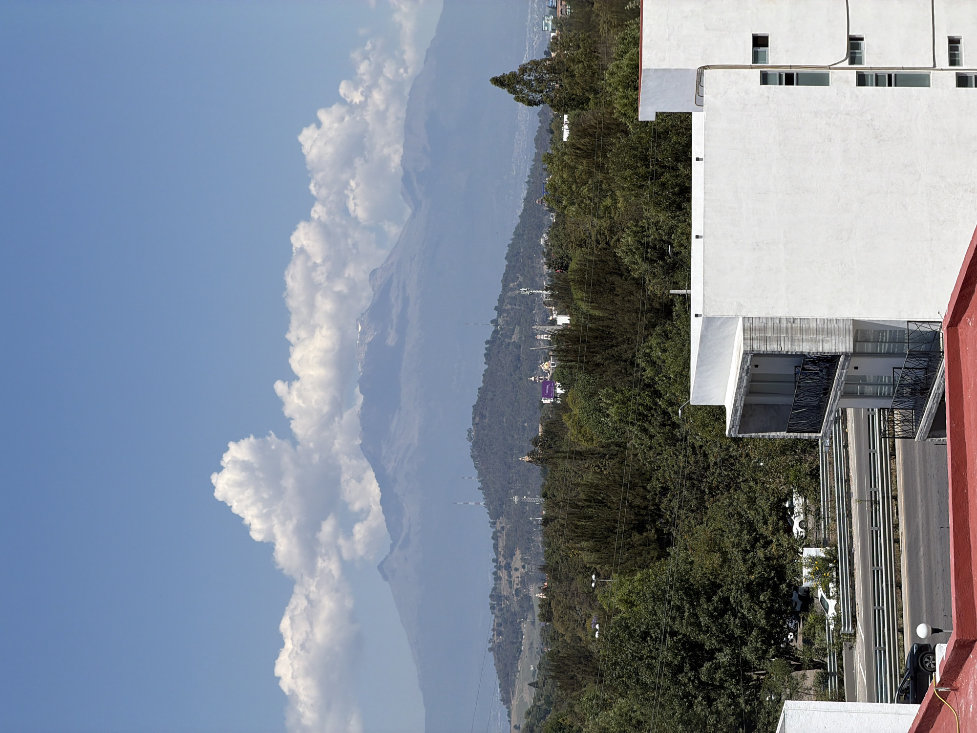 This image presents a panoramic daytime view over San Andrés Cholula, Mexico, capturing a blend of urban elements and natural grandeur. In the foreground on the right, modern white multi-story buildings with various windows and balconies are visible, some featuring exposed utility lines. A reddish-orange wall or rooftop edge runs along the very bottom right and extends towards the foreground. Below the buildings, a street or road is partially visible with a parked black car and what appears to be a white car further down.
The mid-ground is dominated by a dense expanse of green trees, suggesting a hilly or forested area. Nestled within and beyond this greenery, a town or city unfolds, characterized by numerous smaller buildings and houses. Distinct features include a prominent purple billboard with white text, a golden-domed building (possibly a church), and several communication towers or antennas rising from the landscape. Cars can be seen driving or parked on the roads within the town.
In the background, majestic mountains rise, their peaks partially shrouded in atmospheric haze or light clouds. Some hints of snow or bare rock can be seen on the highest visible slopes. The sky above is a clear, bright blue, adorned with scattered, fluffy white cumulus clouds, particularly clustered on the left side of the frame. The weather appears fair and sunny, indicating good visibility despite the distant haze. No specific human activity or event is prominently featured; the image captures a static landscape view from a vantage point, likely from a higher floor of a building.