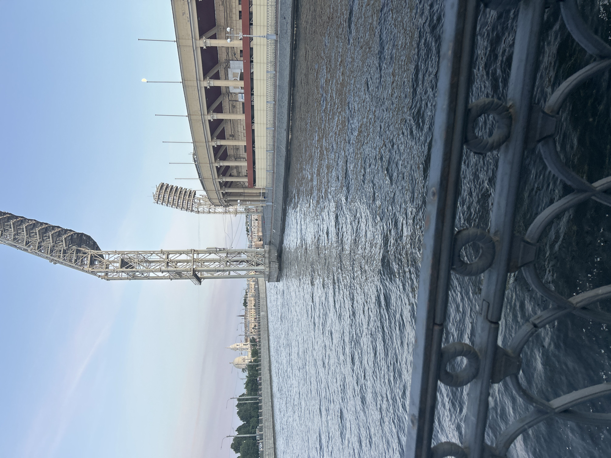 The image shows a view of a body of water next to a concrete embankment and a large stadium structure. A metal railing is in the foreground on the right side of the image. The water is choppy and appears to be a river or canal. On the left side of the image, a large, complex metal structure with many supports and beams extends upwards, possibly a bridge or part of a stadium's lighting rig. Beyond that, a large, tiered stadium is visible, with rows of seats and a supporting structure of columns and beams. The sky is clear and a pale blue, suggesting it is daytime. In the distance, across the water, there is a cityscape with buildings and trees. The overall scene is urban and industrial, with a focus on large architectural structures and the water. The image is oriented vertically, with the top of the stadium and sky on the left.