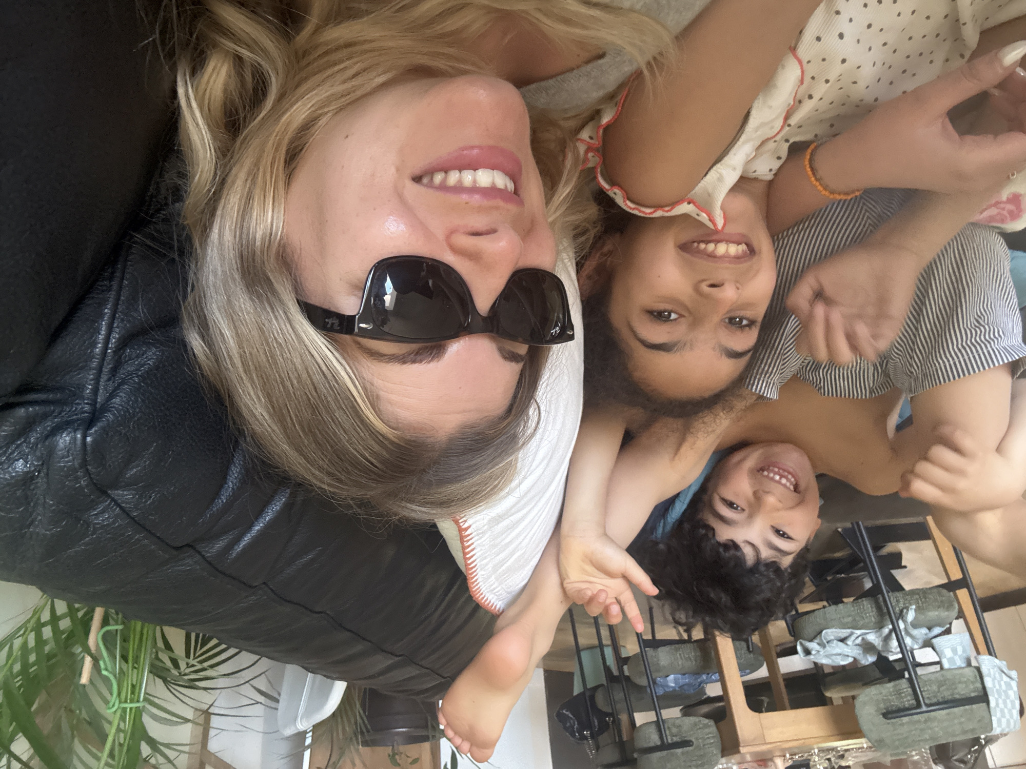 Three people are lying together. One is an adult, a woman with blonde hair wearing sunglasses. Next to her are two children, a girl and a boy. The group is lying on a black leather couch with a white pillow. A green plant is visible in the lower left corner. The location includes a wooden shelf holding multiple green concrete weights. The image was taken in a room in Rousset, France.