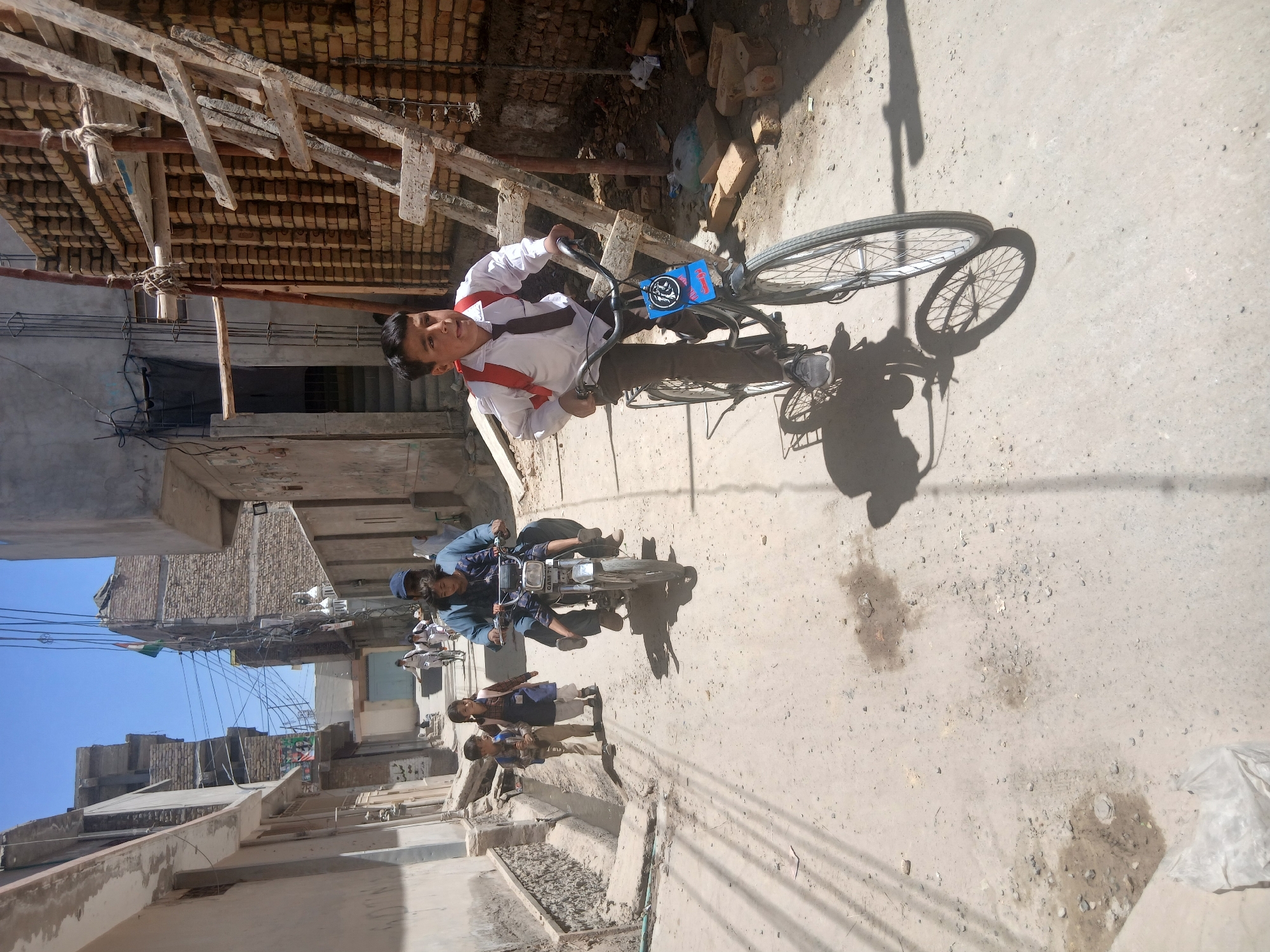 The image shows a street scene in Quetta City Tehsil, Pakistan. A young boy in a school uniform rides a bicycle on a dirt road. Another person rides a motorcycle with children as passengers, and other people walk along the side of the road. Buildings line the street, and there is construction debris visible. The sky is blue.