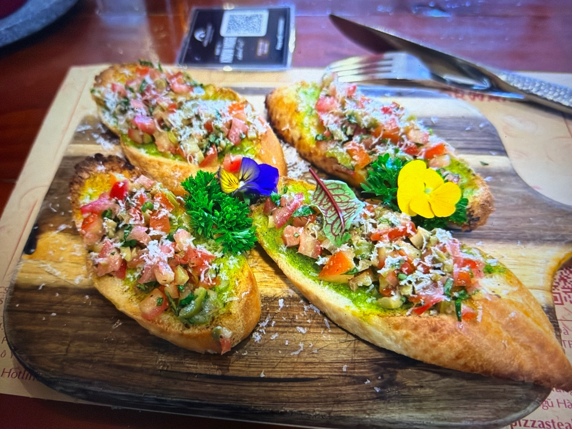 The image shows a close-up of four pieces of bruschetta arranged on a wooden cutting board. The bruschetta are made with toasted baguette slices topped with a green spread, chopped tomatoes, and shredded cheese. Some pieces are garnished with parsley and edible flowers. In the background, a fork and knife are visible, along with a QR code menu. The setting appears to be a restaurant in Da Nang, Vietnam, as suggested by the text on the menu which includes Vietnamese characters and the word "pizzasteak." The lighting is warm and highlights the textures of the food. There are no people or active interactions depicted in the image.