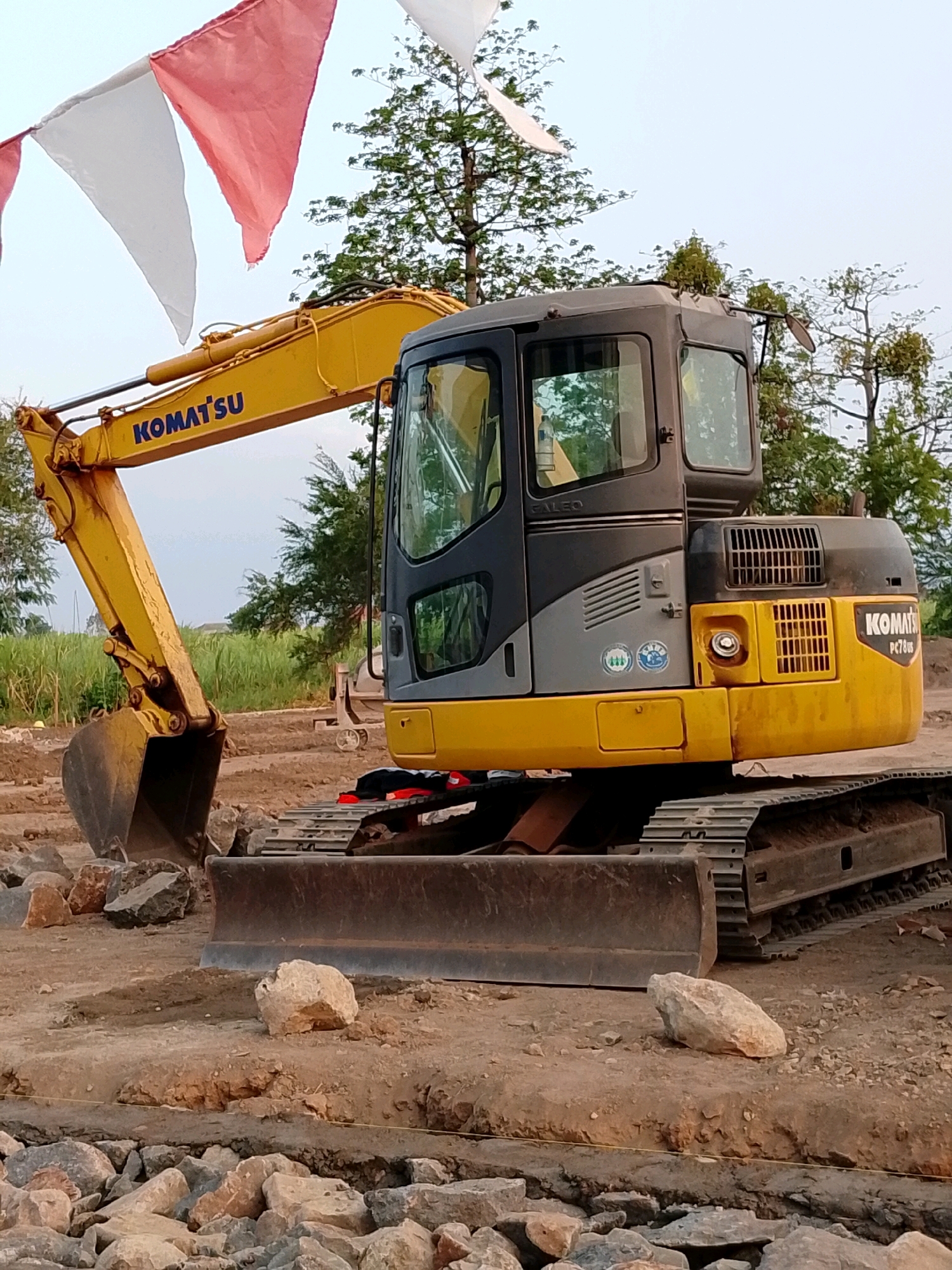 An excavator, identified by the "KOMATSU" logo, is positioned on a dirt construction site. The excavator is yellow and gray, with a visible bucket and tracks. Decorative flags, red and white, are strung above and to the left of the excavator. The background consists of green foliage and trees under a bright sky. The scene appears to be outdoors in a rural or undeveloped area, likely in Pasuruan, Indonesia, given the presence of construction activities and vegetation common to the region.