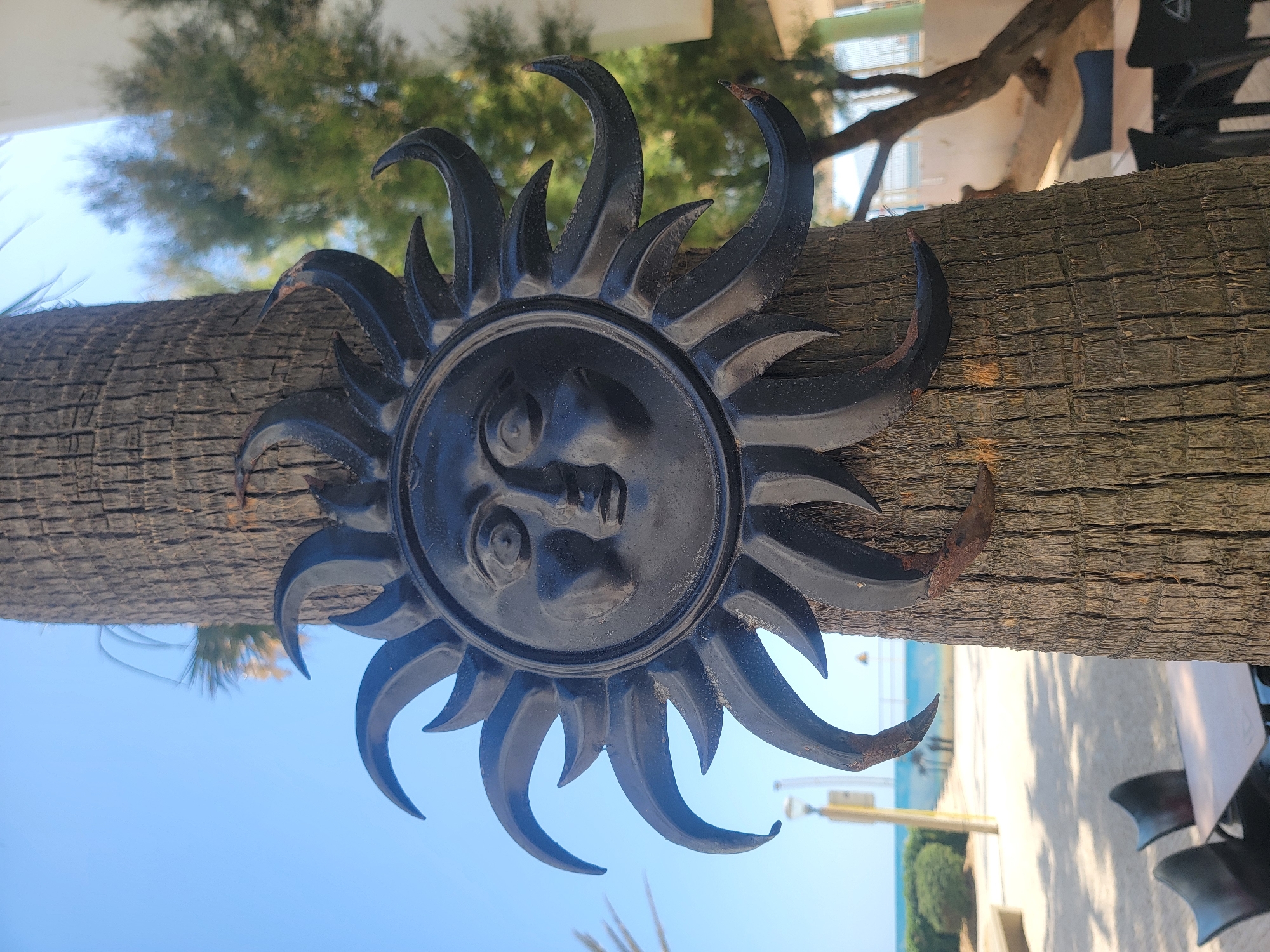 A black metal sun ornament with a face is attached to the trunk of a palm tree. The ornament has a circular center with a sculpted face and many pointed rays radiating outward. The palm tree trunk is rough and textured, with a dark brown bark. The background is blurred, showing a beach scene with a blue sky, the ocean, some greenery, a building, and outdoor seating with chairs and tables. The overall impression is a sunny day, likely in a coastal area. This image was captured in Quarteira, Portugal, given the characteristic palm trees and coastal setting of the city.