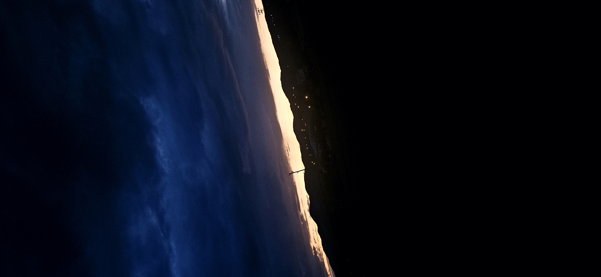 This landscape photograph captures a scene at either dusk or dawn over a hilly or mountainous region in Momin Prohod, Bulgaria. The image is composed with a clear division between the sky, a luminous horizon, and a dark landmass.
The upper portion of the frame features a deep, dark blue sky, transitioning slightly to lighter shades where subtle, wispy cloud formations are visible. A prominent, glowing band of light, colored in warm yellows and oranges, stretches horizontally across the middle of the image, signifying the setting or rising sun.
Below this radiant horizon, the landmass is rendered in a dark silhouette, suggesting rolling hills or mountains. Scattered across this dark terrain are numerous small, bright artificial lights, indicating the presence of a distant town or scattered buildings. Two tall, slender utility poles, possibly power line towers, are visible in silhouette against the bright horizon, hinting at infrastructure.
The lower-right quadrant of the image is predominantly black, either representing an extremely dark foreground that absorbed all available light or an intentional framing choice.
No people or specific activities are depicted, and no visible text is present. The overall mood is serene and atmospheric, emphasizing the natural beauty of the transition between night and day.