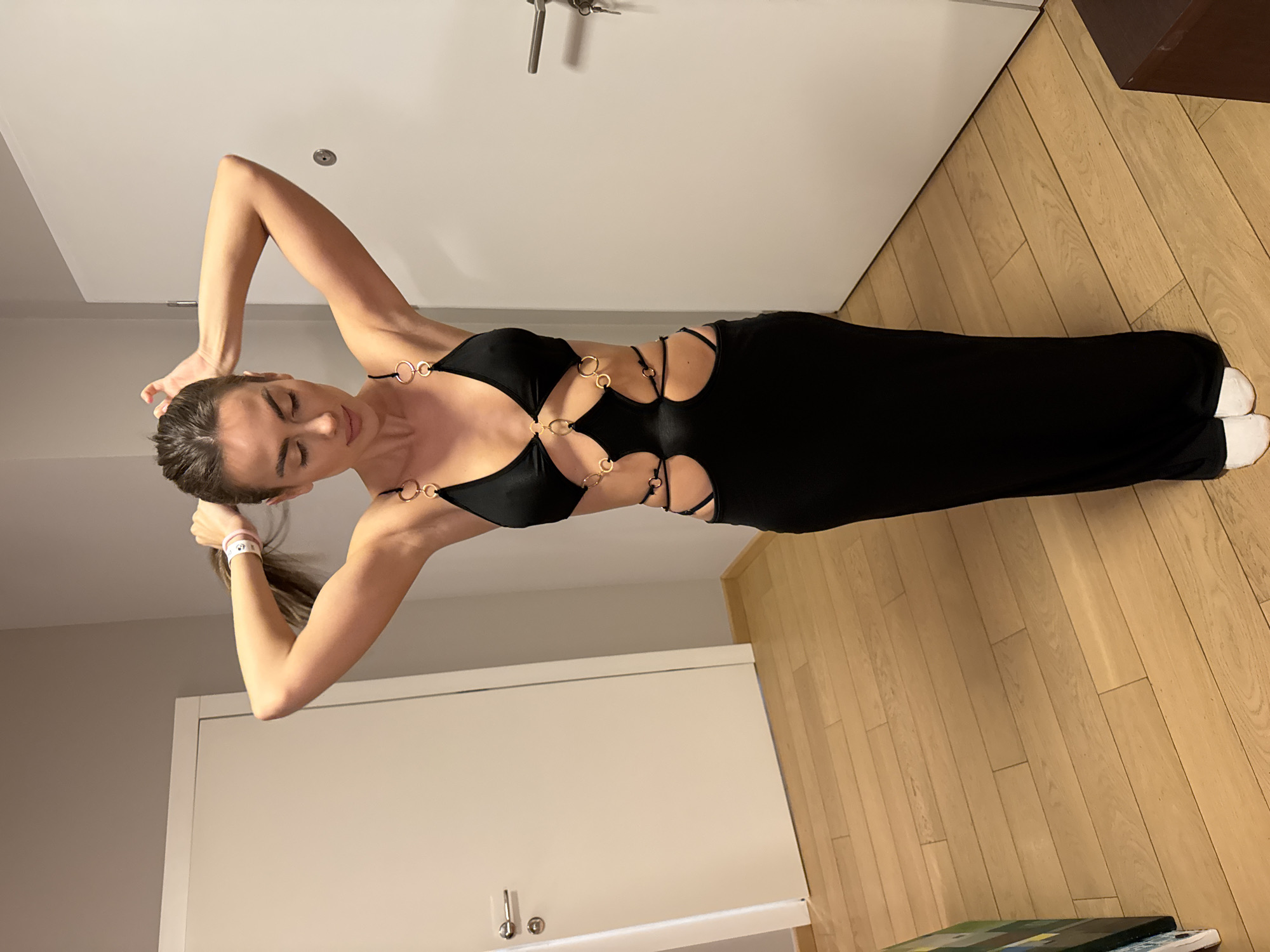 A woman is standing indoors, facing slightly to the left, and looking down. She is wearing a black dress with circular metal accents around the chest and torso cutouts. Her arms are raised above her head, and she is holding her hair in a ponytail with a white wristband around it. She has fair skin and dark hair. She is wearing white socks.
The location appears to be inside a residential space, possibly a hotel or apartment room in Warsaw, Poland. The floor is hardwood with a light-colored finish, and a portion of the floor and a decorative object are visible in the bottom right corner. The walls are painted in a neutral color, and there are white doors visible in the background, some with silver door handles. A dark wooden cabinet is visible in the upper right corner.