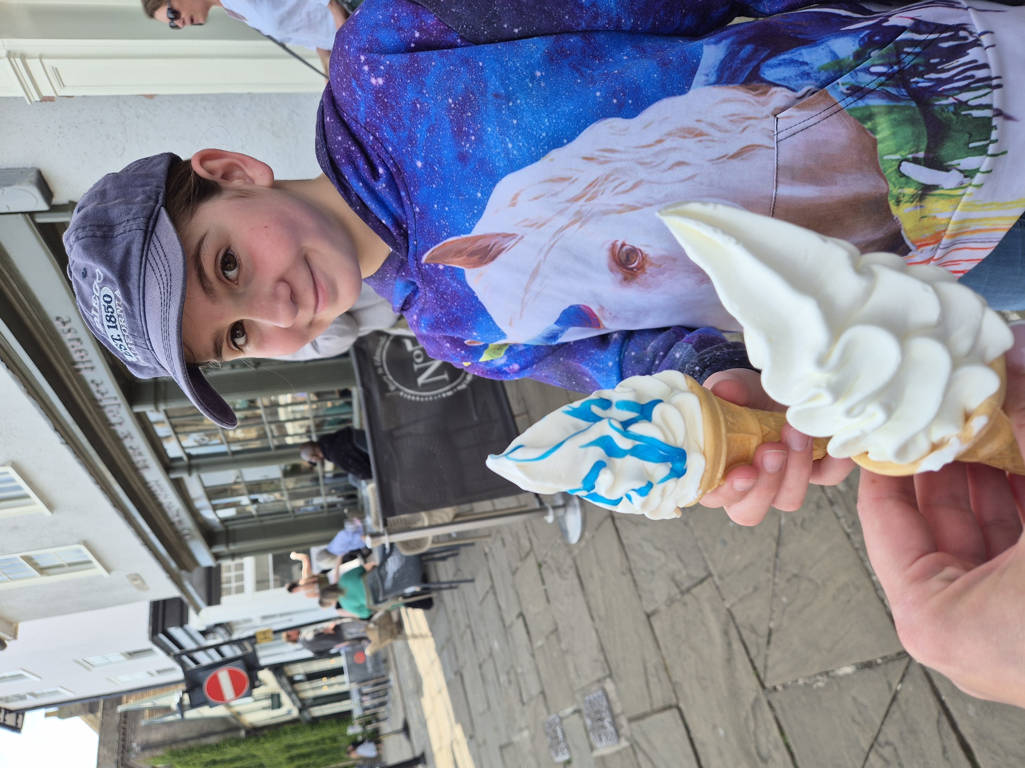 A photograph captured in the city of West Suffolk, United Kingdom, shows a child in the upper left of the frame wearing a gray baseball cap and a blue and purple hooded sweatshirt featuring an image of a white horse. The child is holding two ice cream cones. One cone has white soft-serve ice cream with blue swirls, while the other has plain white soft-serve. An arm and hand appear in the lower right, holding the plain white ice cream cone. In the background, a street scene unfolds with pedestrians, outdoor seating for a cafe or restaurant, buildings, and a traffic sign. The ground is paved with stone.