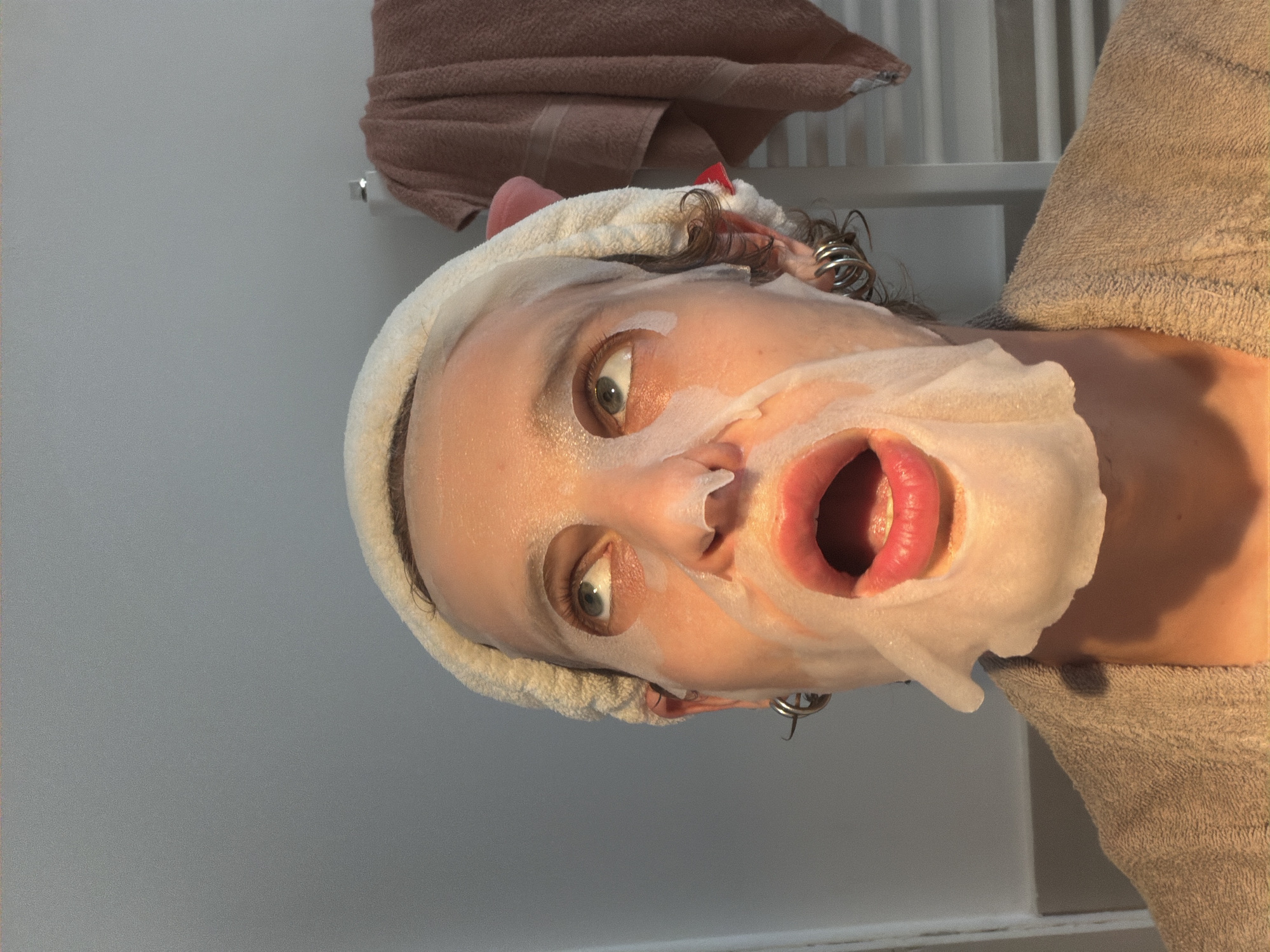 A woman wearing a face mask and a towel on her head is looking directly at the camera with her mouth open. She is wearing a robe and there is a towel rack behind her. There is no indication of the location being in the city of Milan, Italy.