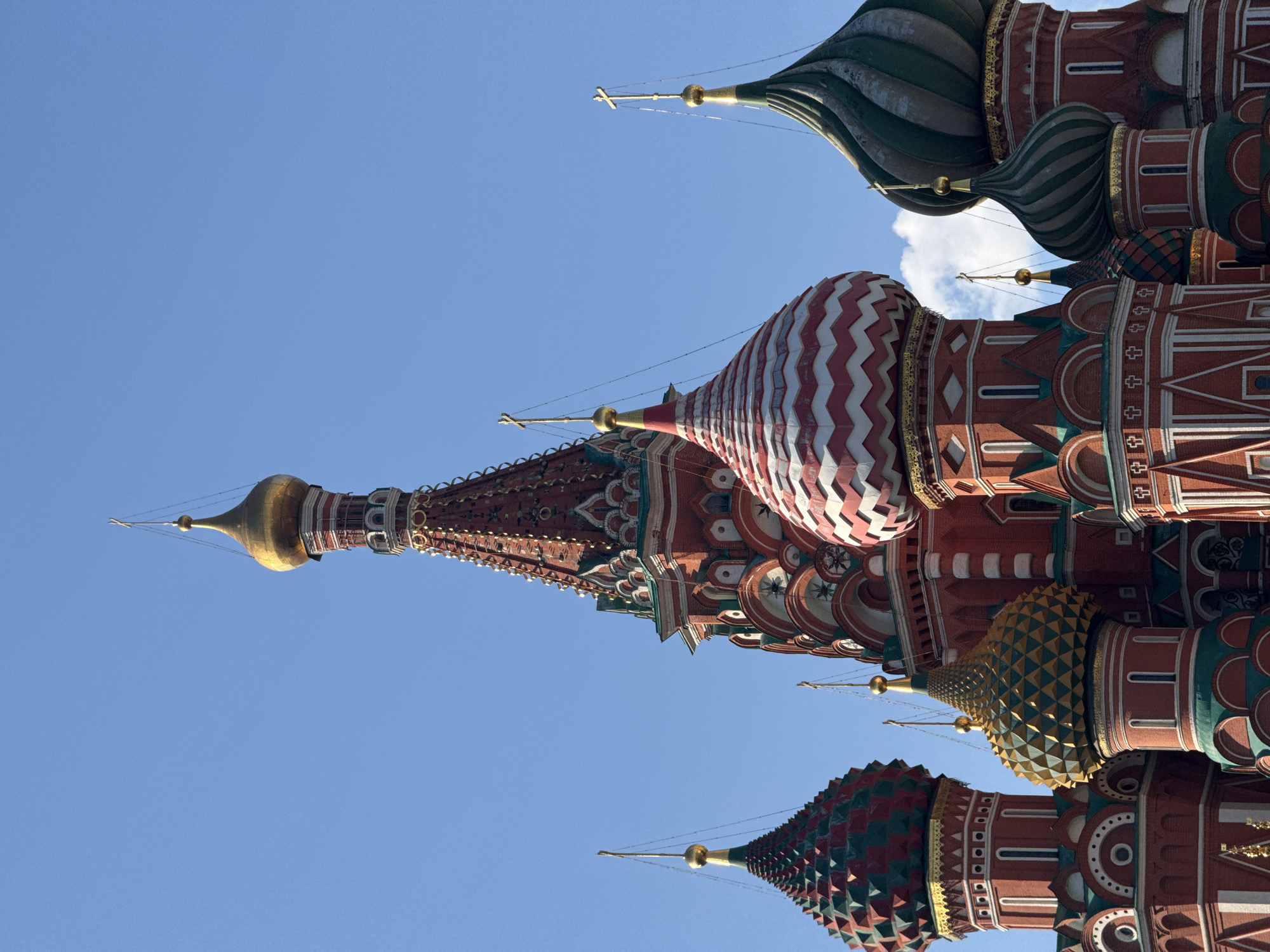 The image shows a close-up, upward-angled view of the domes of St. Basil's Cathedral in Moscow, Russia. The sky is a clear, bright blue. The domes are elaborately decorated with varied colors and patterns, including stripes, diamond shapes, and geometric motifs. Each dome is topped with a golden cross. The architecture is of Russian Orthodox style.